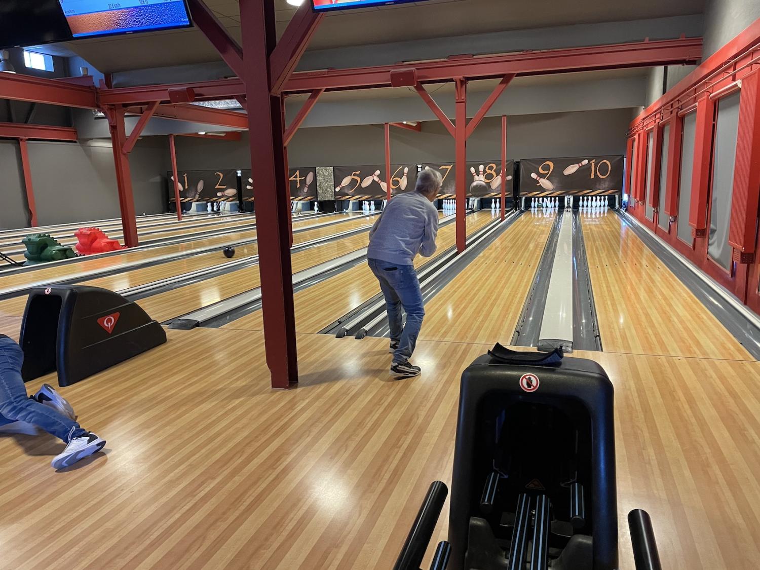 Person som bowlar i en inomhusgränd med flera banor, iklädd ledig klädsel. Andra människor syns i bakgrunden. Bowlingklot finns på ställ i närheten.