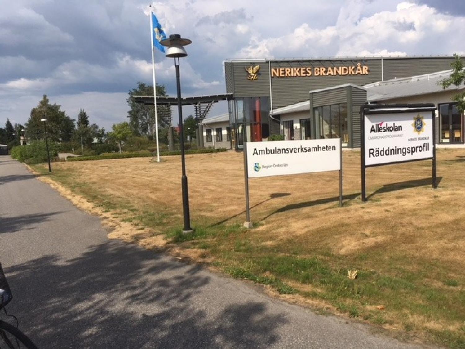 En byggnad med skyltar där det står ”Ambulansverksamheten” och ”Räddningsprofil” står på en gräsmatta nära en väg, med en flaggstång och en molnig himmel i bakgrunden.