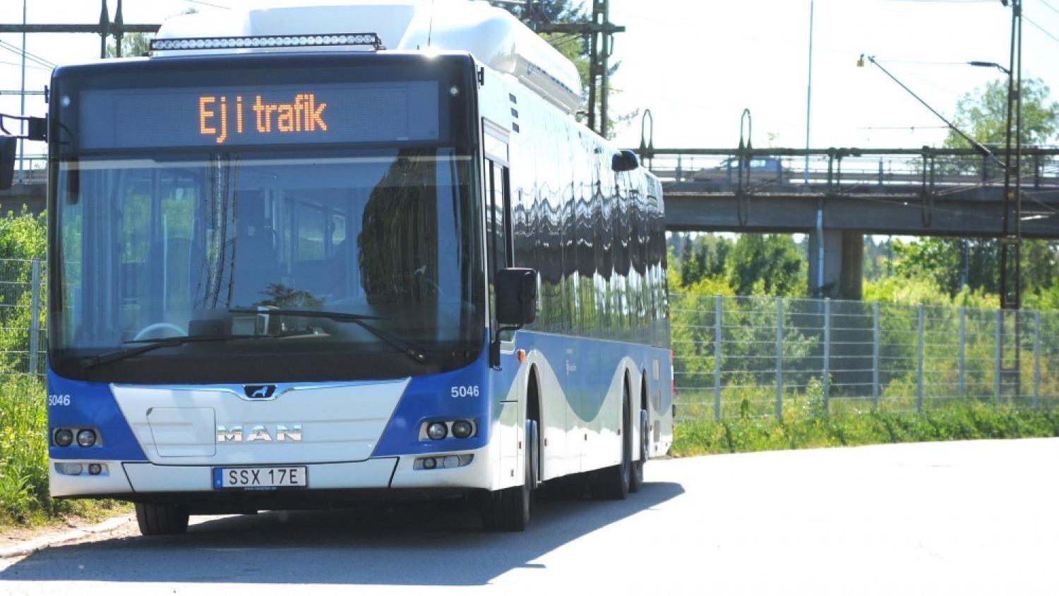 En blå och vit MAN-buss med skylten ”Ej i trafik” stod parkerad på en solbelyst gata nära ett staket och en viadukt.