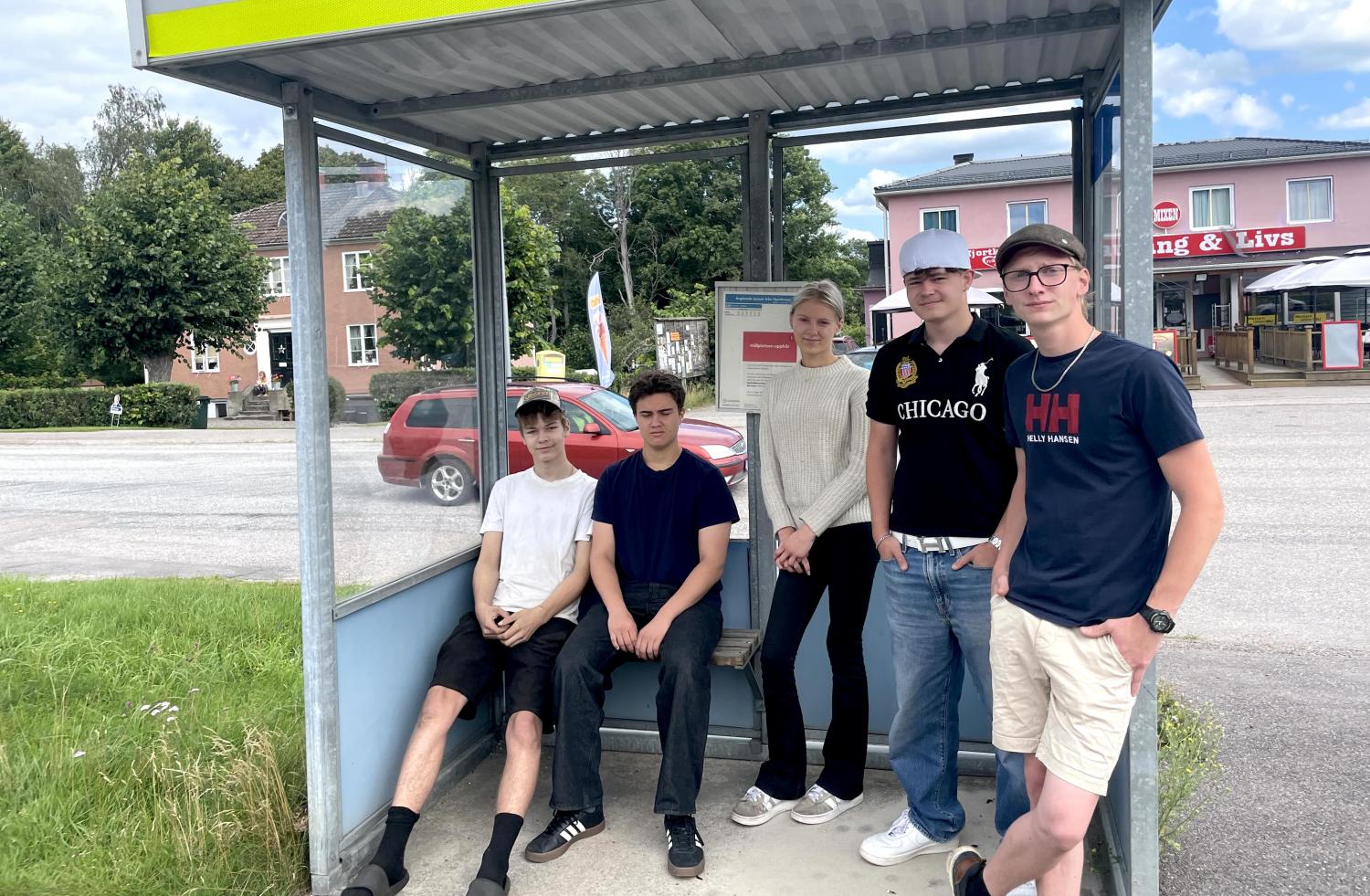 Fem unga människor står och sitter vid en busshållplats under tak en solig dag, med byggnader, bilar och träd i bakgrunden.