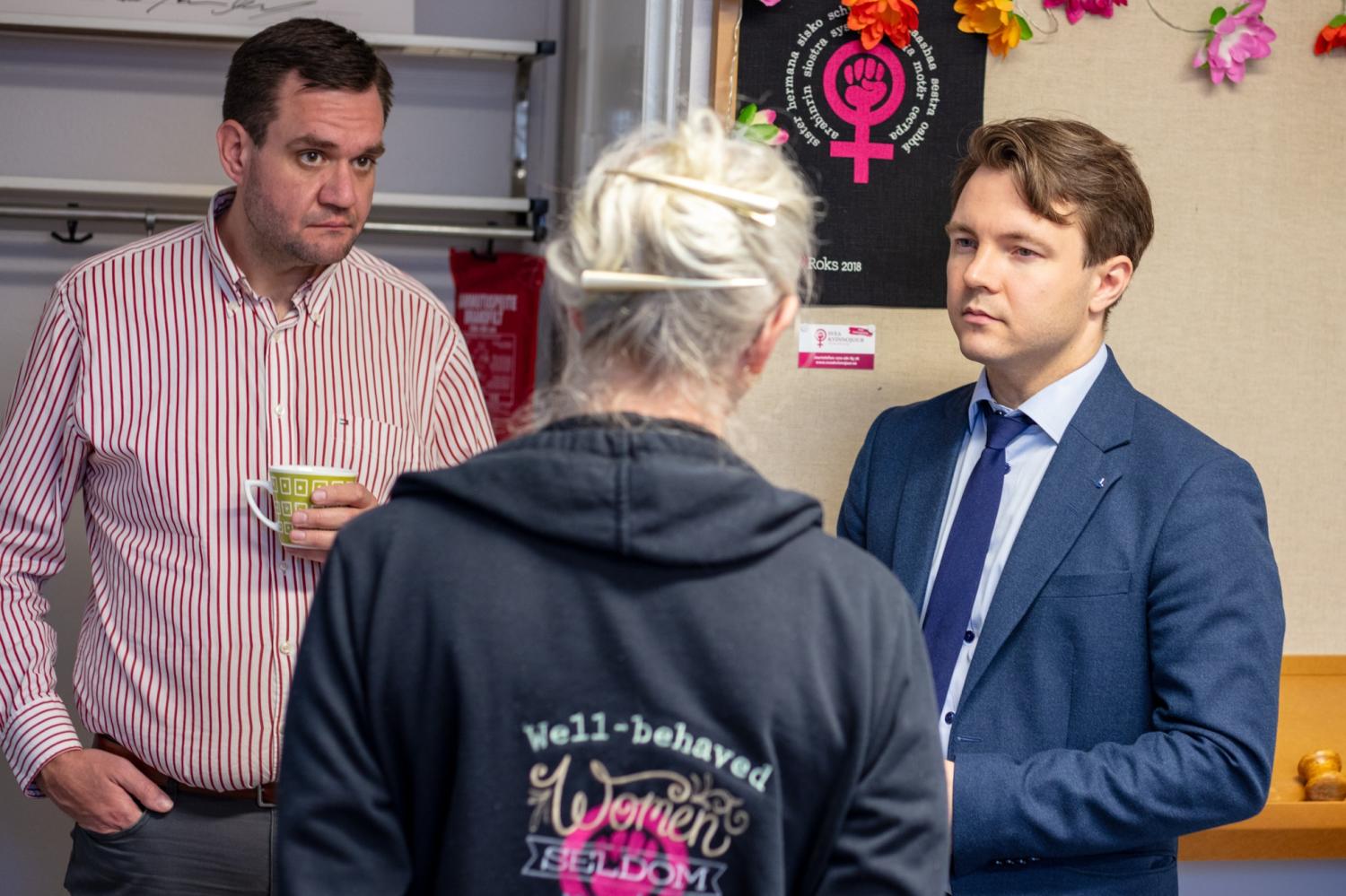 Tre personer samtalar inomhus; en man håller i en mugg, en annan är klädd i kostym och en person som står med ryggen mot kameran bär en huvtröja med en slogan.