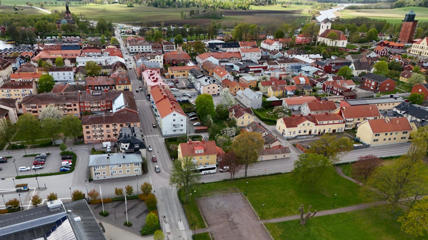 Flygfoto över en liten stad med röda tak, trädkantade gator och öppna grönområden omgivna av åkrar och jordbruksmark.
