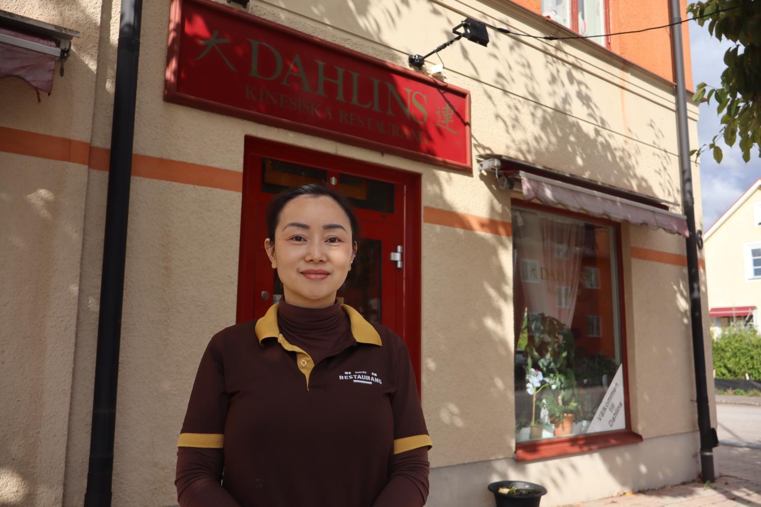 En kvinna i brun uniform står framför en beige byggnad med en röd skylt där det står ”Dahlins Kinesiska Restaurang”.