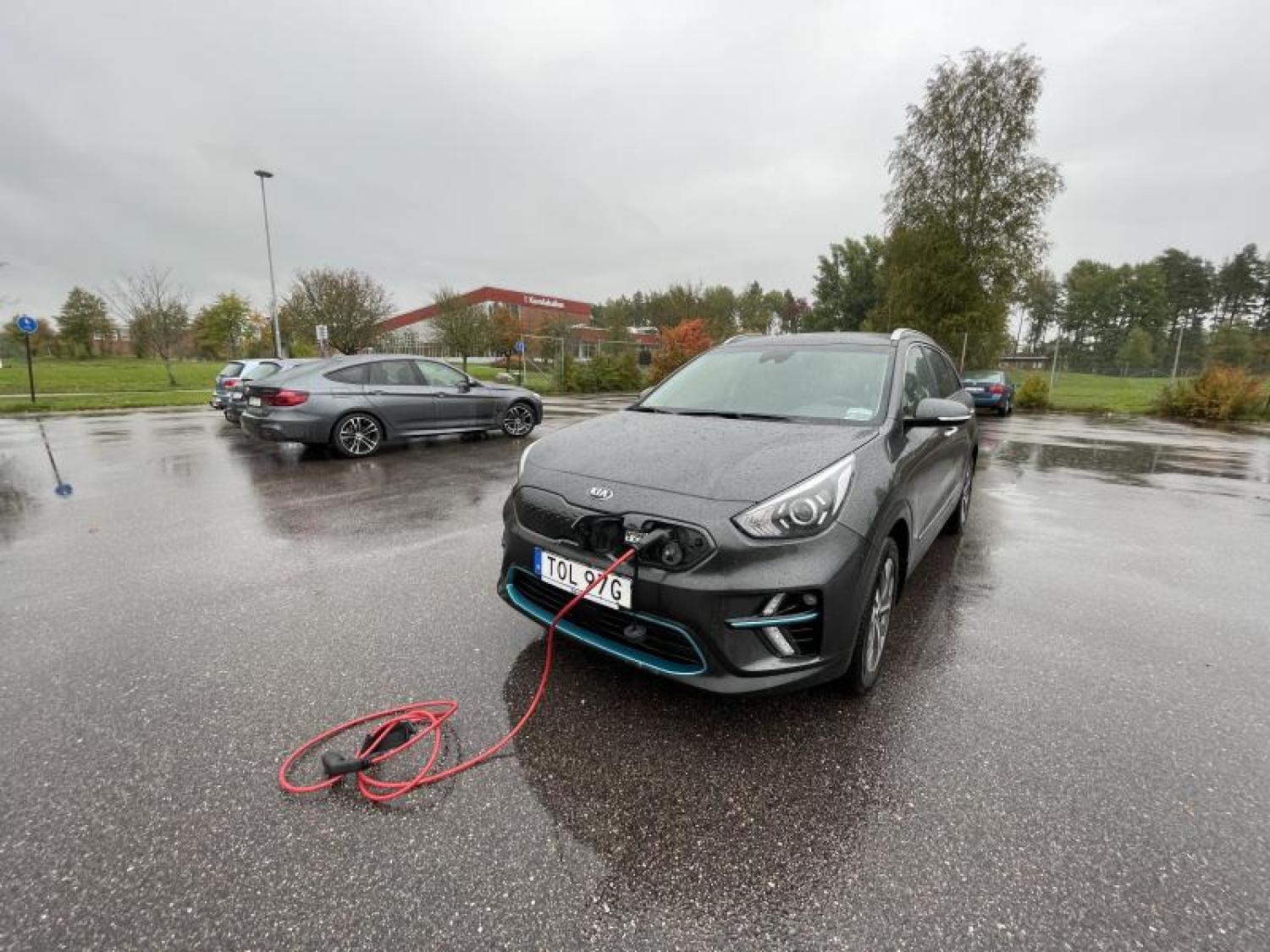En grå elbil står parkerad på en våt parkeringsplats och laddas via en röd kabel som är ansluten till dess främre port. I bakgrunden syns en annan bil och träd under en regnig dag.