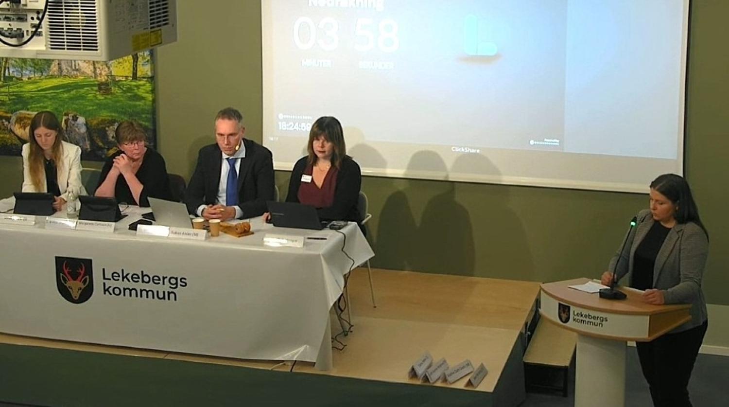 Fem personer befinner sig i ett mötesrum; fyra sitter vid ett bord med bärbara datorer, och en står vid ett talarstativ och talar. På en stor skärm bakom syns en nedräkningsklocka. Platsen är märkt med ”Lekebergs kommun”.