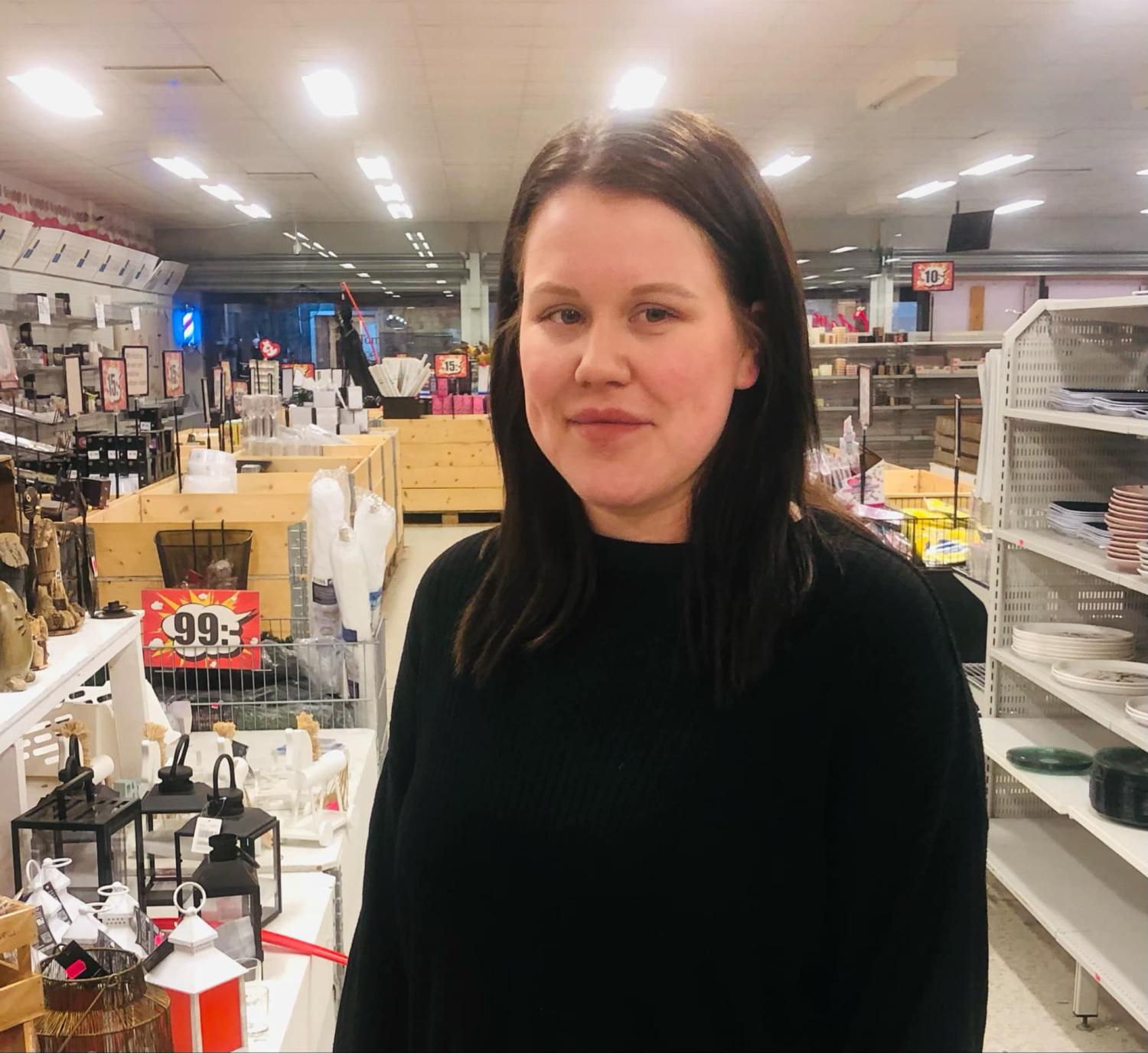 En person med långt mörkt hår står inne i en butik med hyllor och olika hemvaror i bakgrunden.