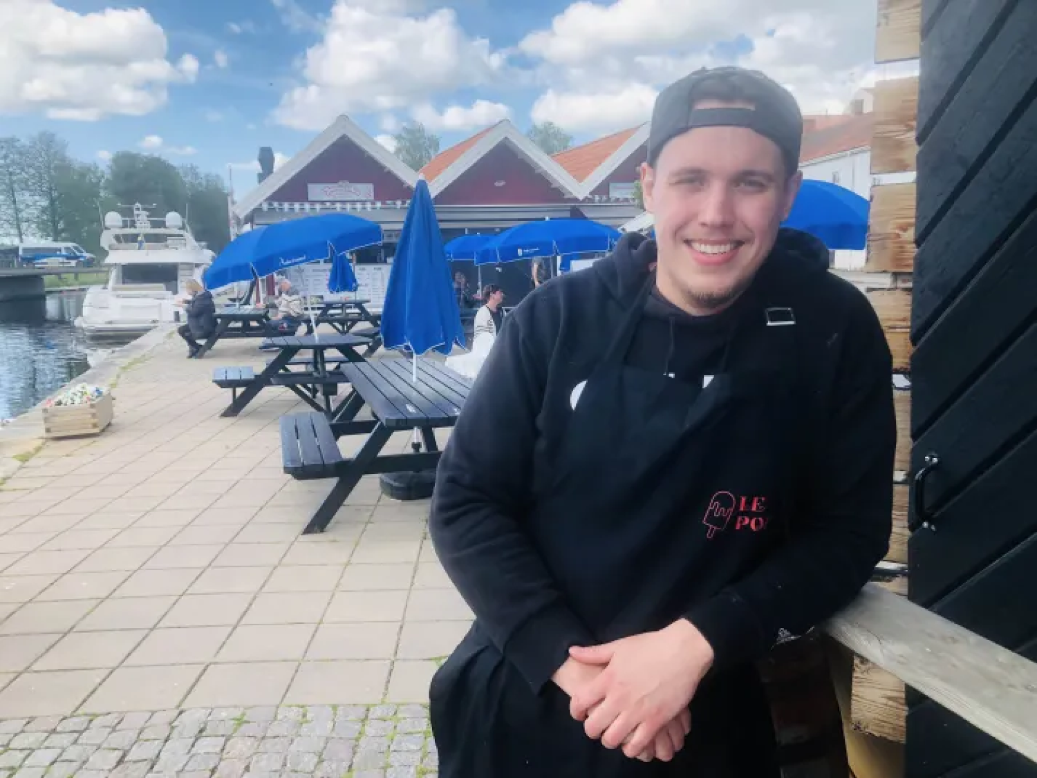 En ung man i svart luvtröja och förkläde ler medan han står utomhus nära picknickbord, blå parasoller och en båt vid kaj en molnig dag.
