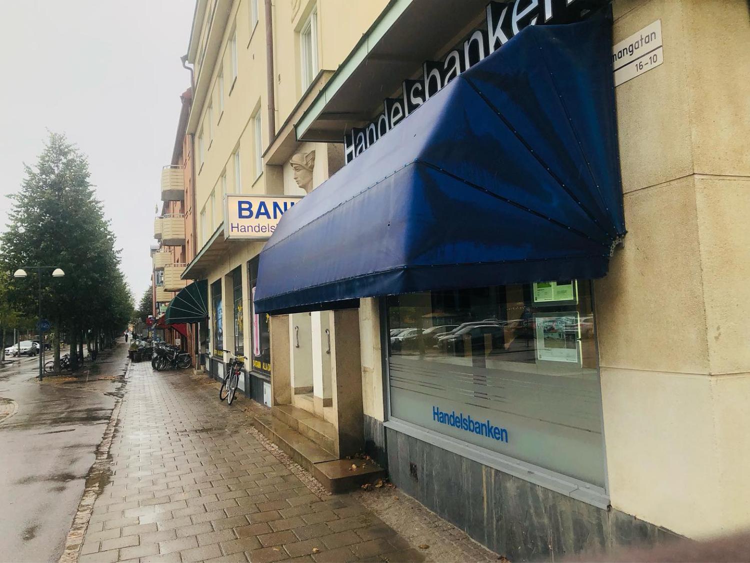 Gatuvy med Handelsbankens kontor med blå markis och närliggande skyltfönster. Cyklar står parkerade längs trottoaren och träd kantar den blöta gatan en molnig dag.