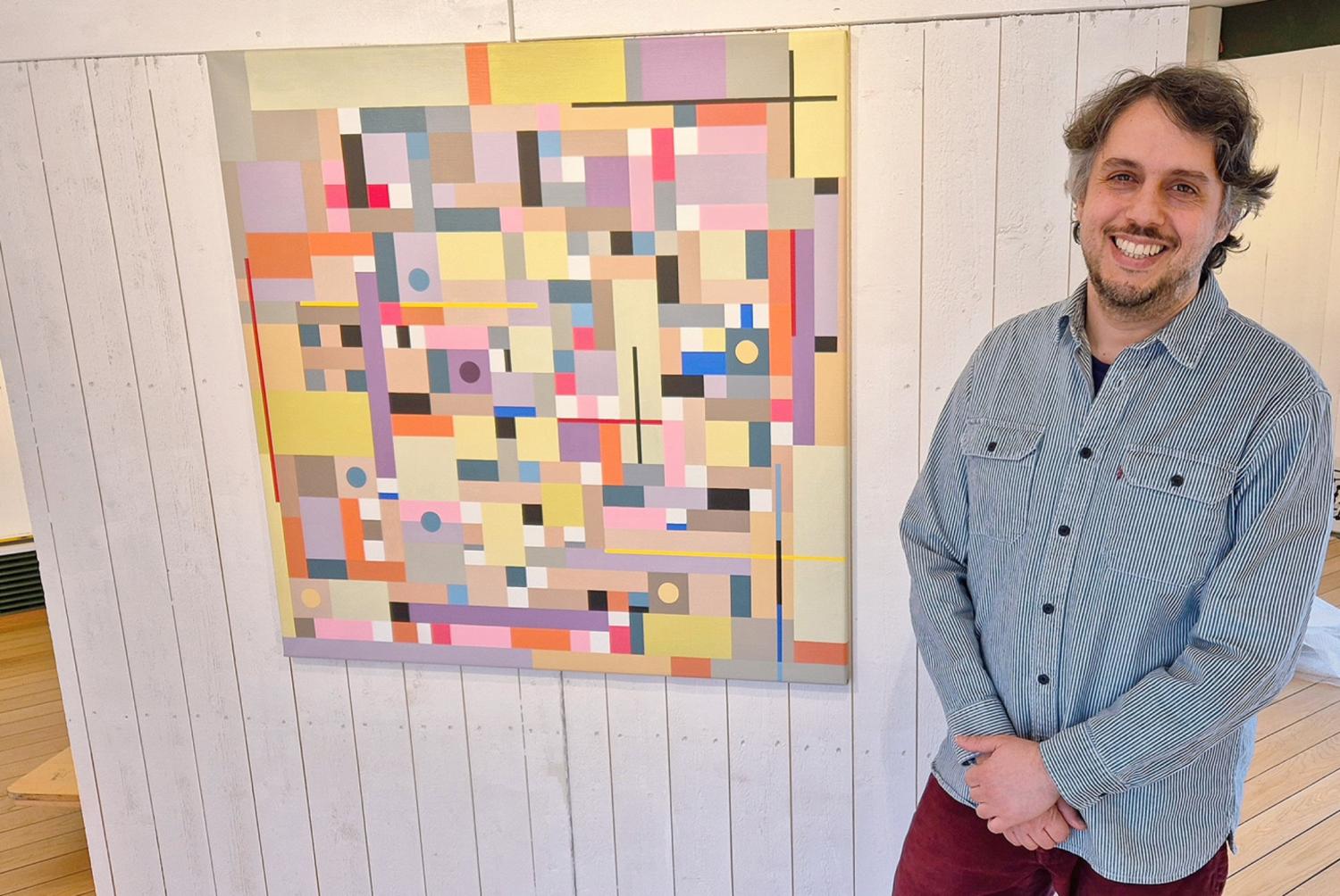 En man står och ler bredvid en färgglad geometrisk abstrakt målning på en vit trävägg.