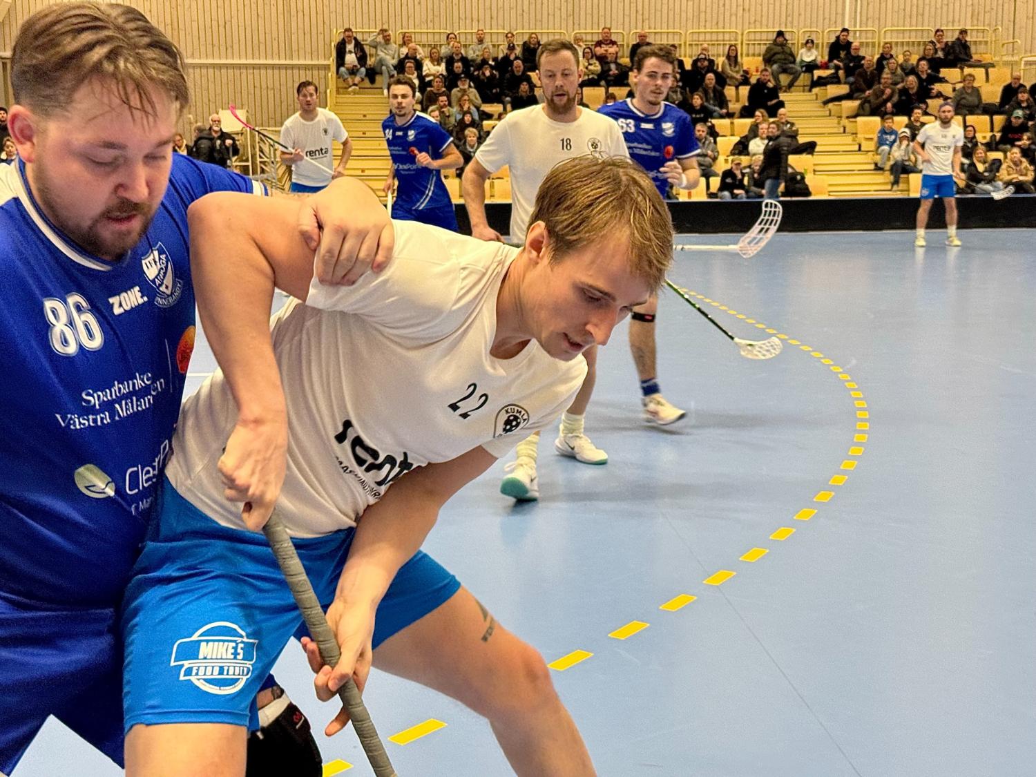 Två innebandyspelare tävlar om kontrollen över bollen nära rinkgränsen, med andra spelare och åskådare synliga i bakgrunden.