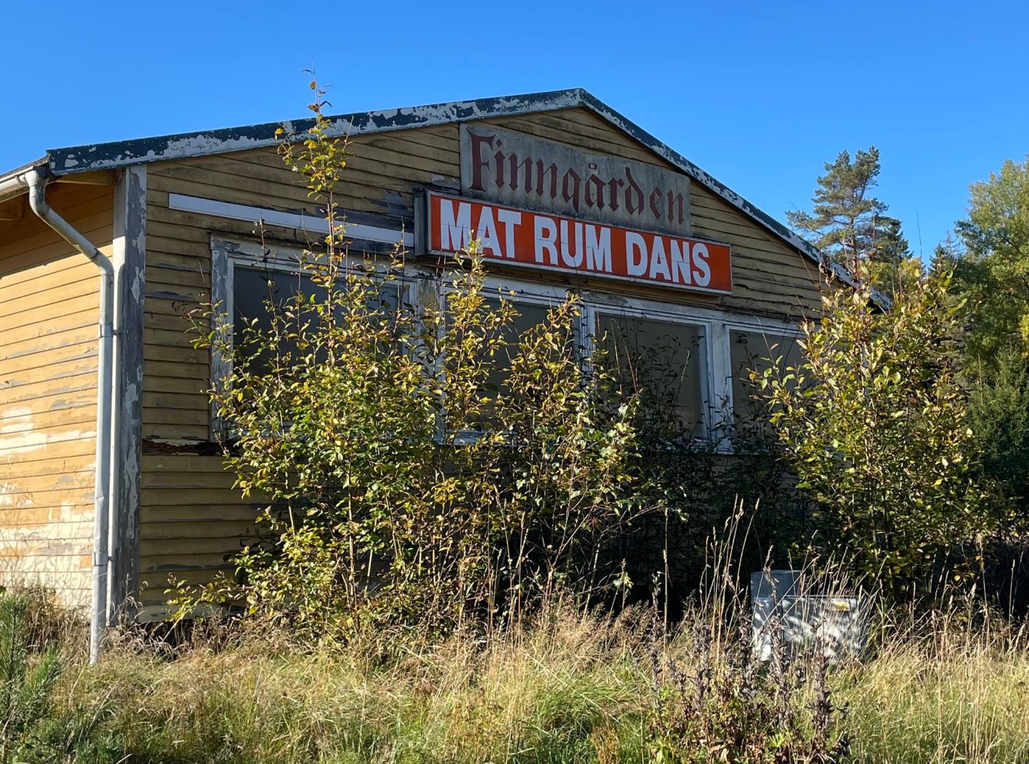 En väderbiten gul träbyggnad med förvuxna buskar framför, med en skylt på svenska som säger