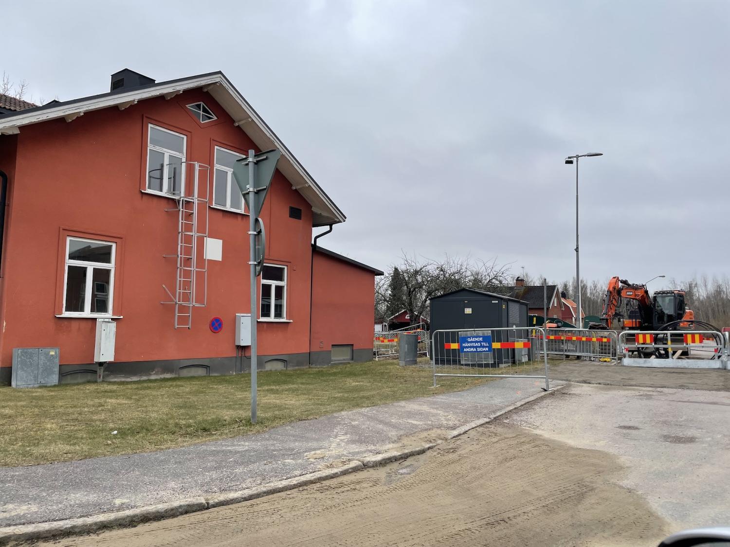 Röd byggnad med metallstege på sidan, i anslutning till vägarbeten med byggbarriärer och grävmaskin. Mulen himmel och gräsyta i förgrunden.