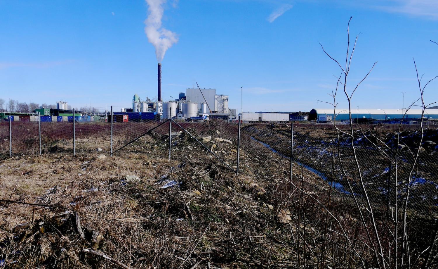 En industrianläggning med rök som stiger upp från en skorsten, sedd bakom ett kedjelänksstängsel med igenvuxet gräs och karg mark i förgrunden. Behållare och lagringstankar är synliga.