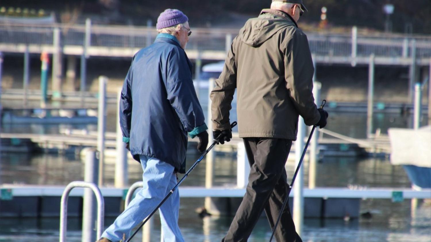 Två äldre personer går sida vid sida nära en småbåtshamn, båda med varsin käpp i handen, klädda i jackor och mössor en klar dag.