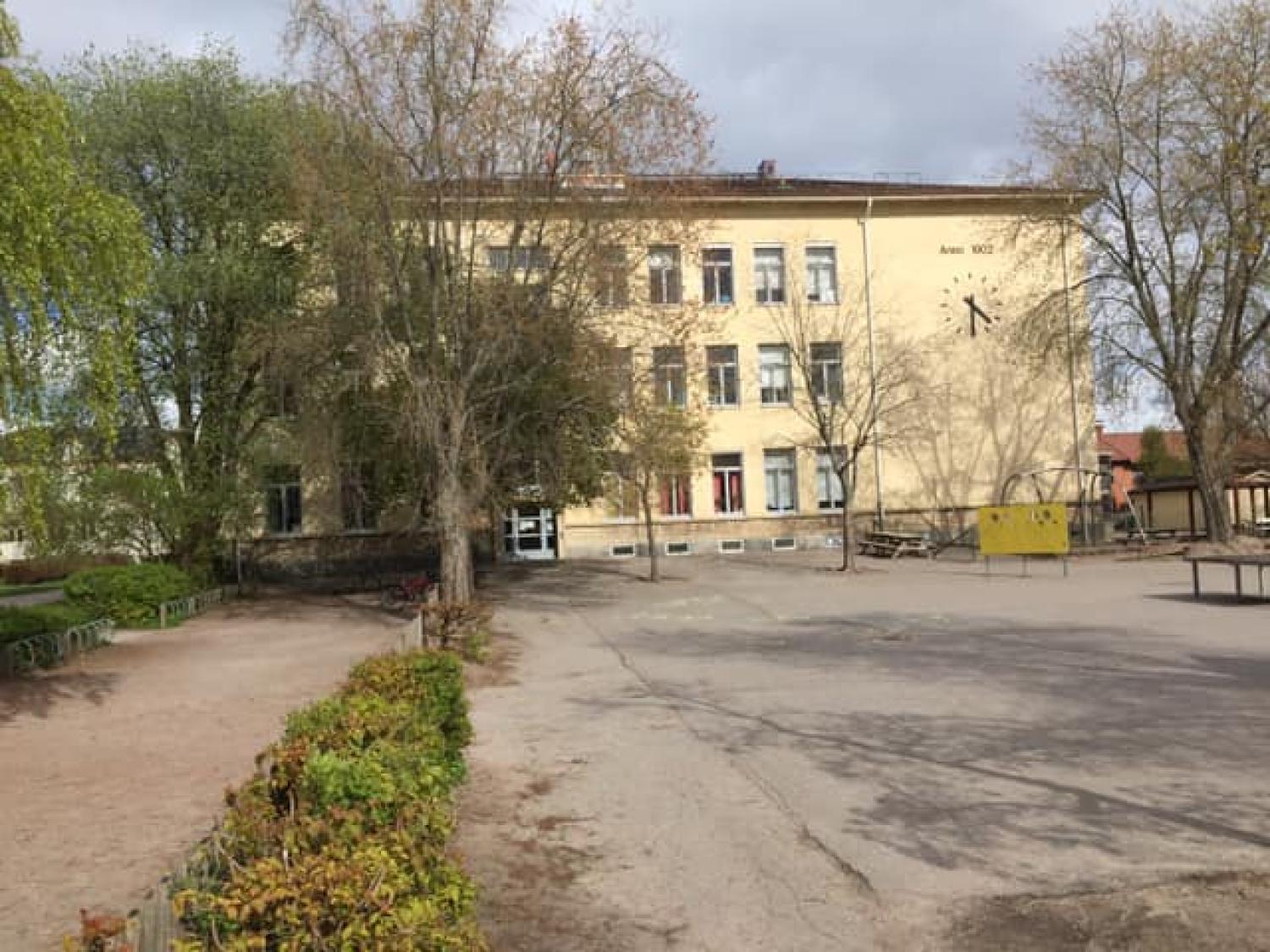 En beige byggnad i tre våningar med en klocka, omgiven av kala träd och en tom stenlagd yta framför. Mulen himmel.