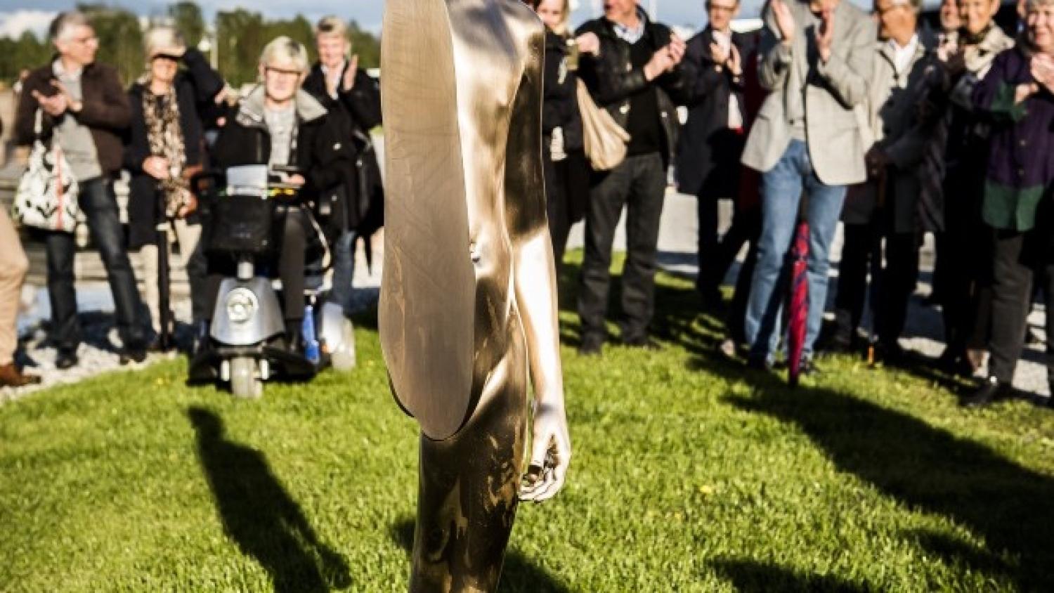 En humanoid skulptur av metall står på gräset medan en grupp människor, varav några klappar händerna, tittar på i bakgrunden.