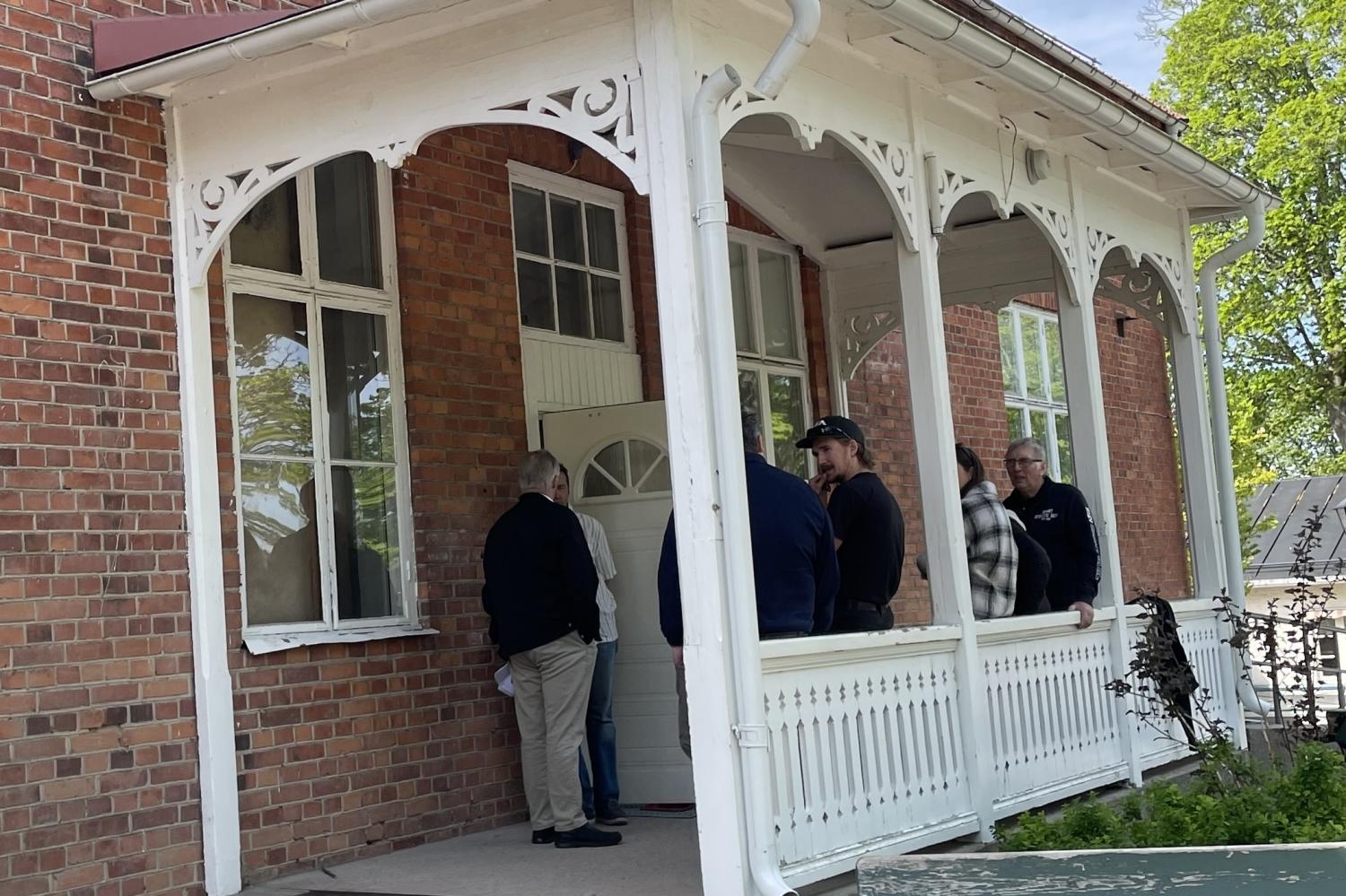 Flera personer står och pratar på en täckt veranda till en röd tegelbyggnad med vita lister och dekorativa träslöjd.