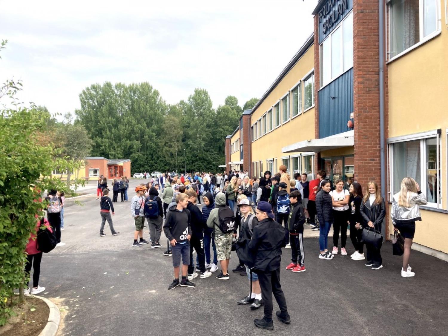 Elever samlas utanför en skolbyggnad och väntar i en utspridd kö, några i samtal, i en förortsmiljö.
