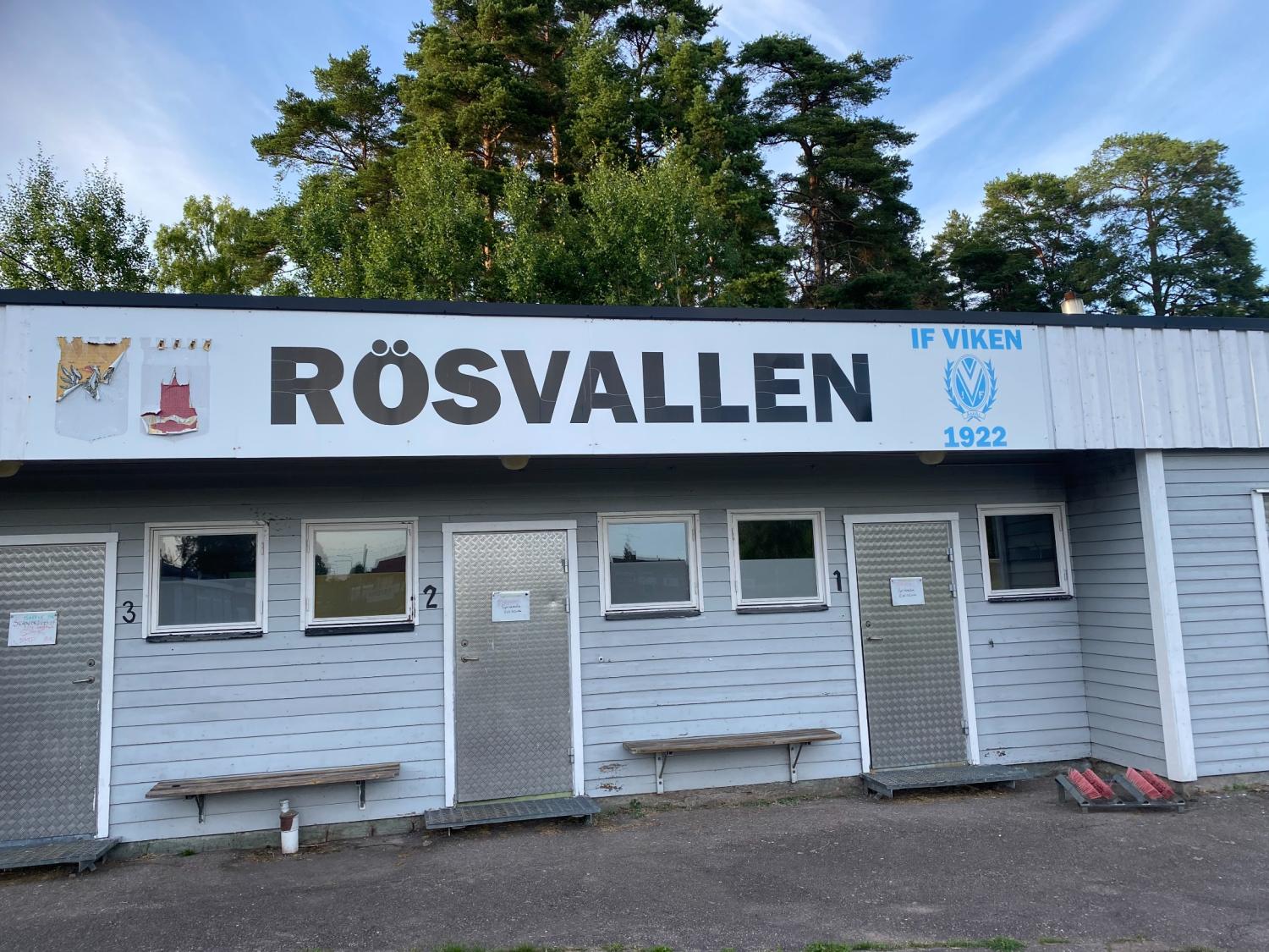 Idrottsanläggningen Rösvallen med numrerade dörrar, en bänk och skyltar, belägen under träden mot en blå himmel.