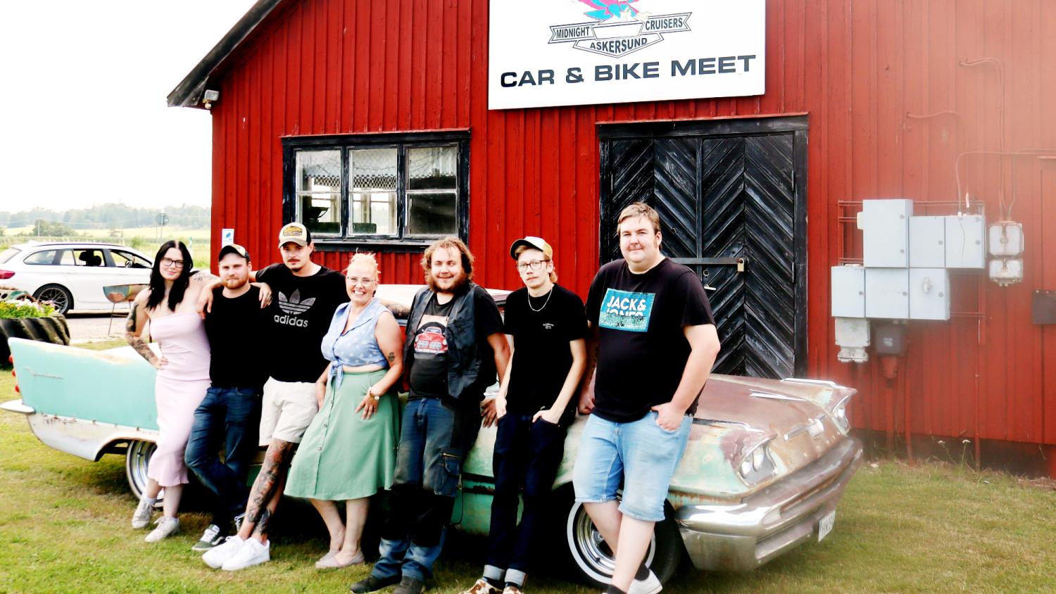 Sju personer står tillsammans framför en veteranbil och en röd byggnad med en skylt där det står ”Car & Bike Meet”.