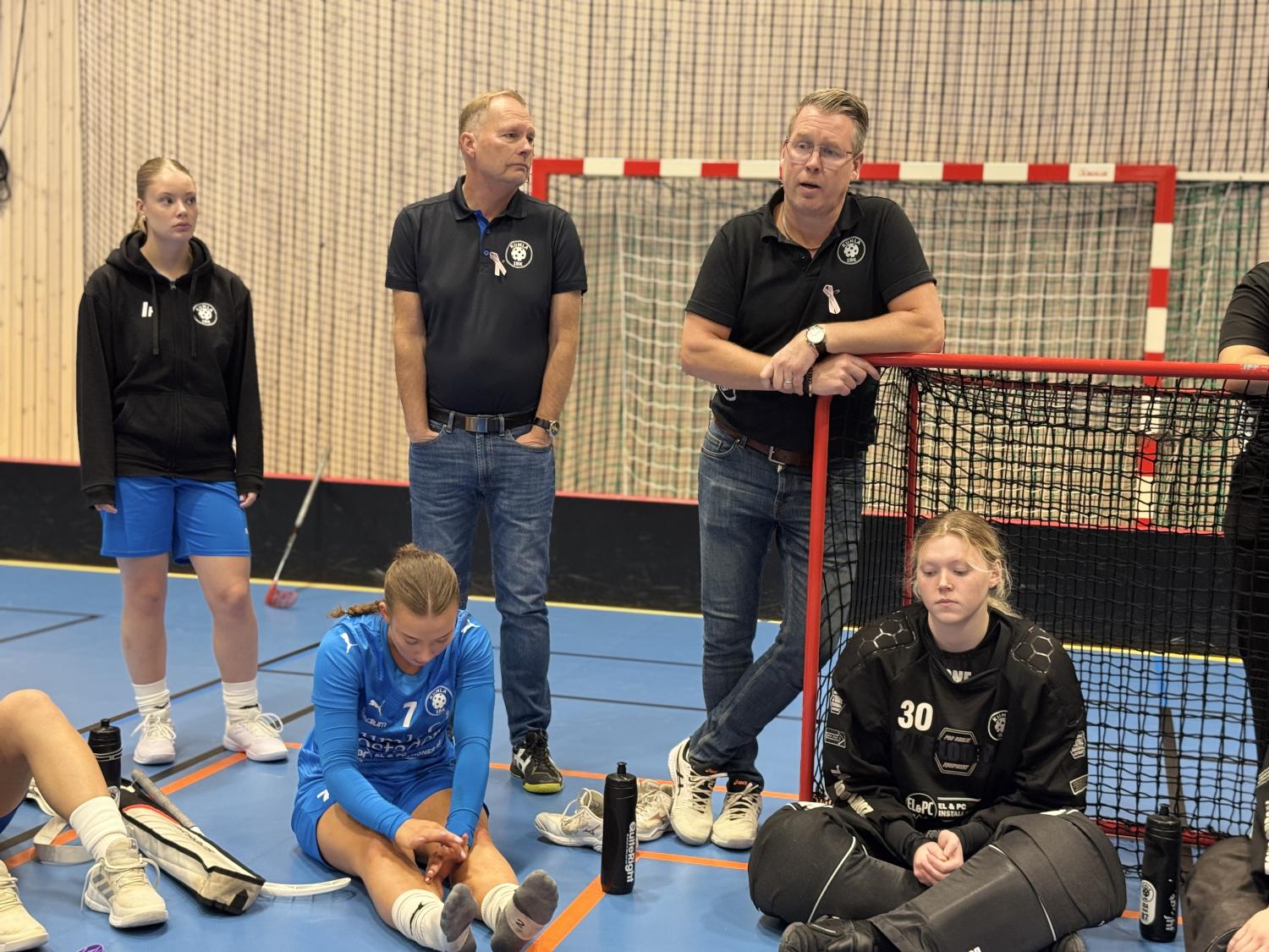 Två tränare talar till ett flicklag i innebandy i en gymnastiksal; några spelare sitter på golvet, varav en i målvaktskläder, medan andra står och lyssnar.
