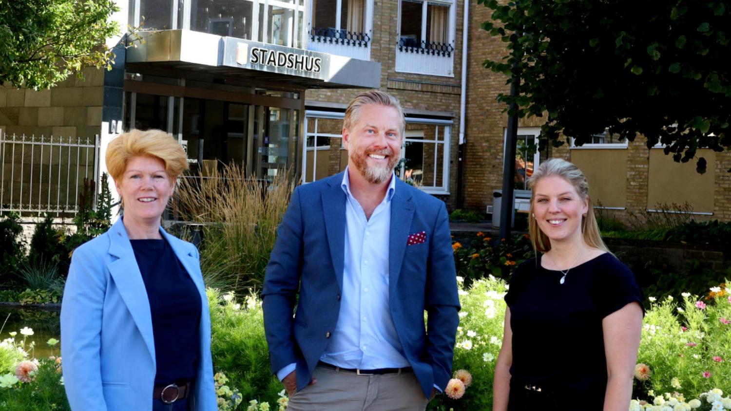 Tre personer i affärskläder står utanför en byggnad med skylten ”STADSHUS”, med blommor och grönska i förgrunden.