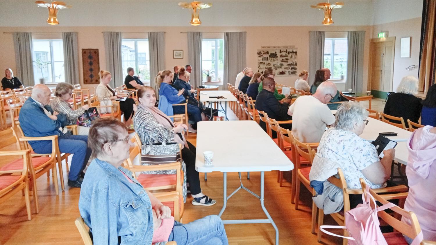 En grupp vuxna sitter på trästolar och vid bord i ett samlingsrum, vända mot framsidan där någon verkar hålla ett tal eller göra en presentation.