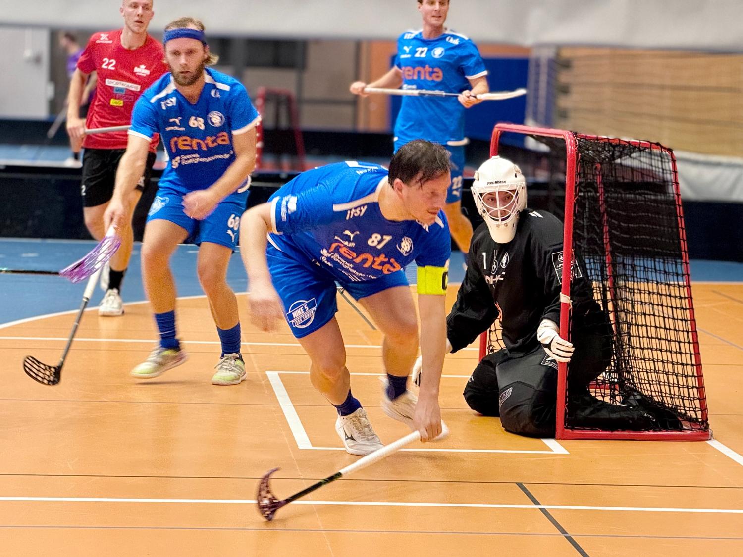 Tre innebandyspelare i blå tröjor springer nära ett mål medan en målvakt i svart knäböjer inne i målet. Spelarna verkar vara i rörelse på en inomhusplan.