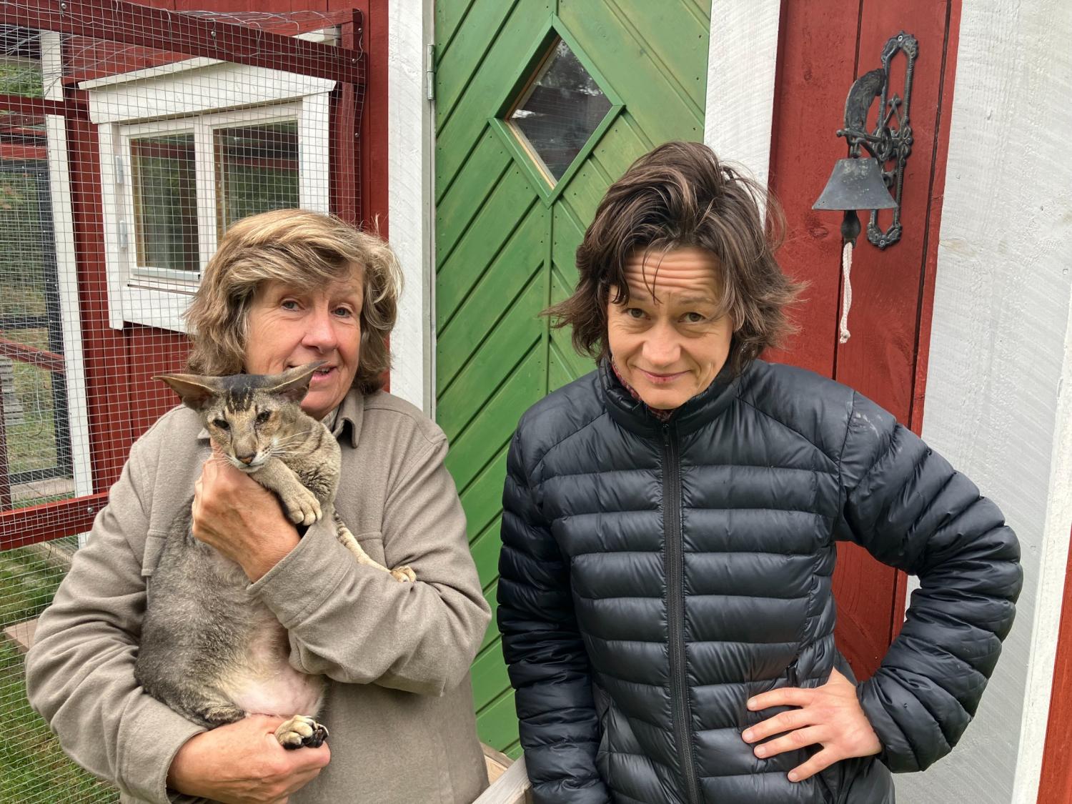 Två personer står utanför en röd och vit byggnad; den ena håller en katt, och den andra står med en hand på höften. I bakgrunden syns en grön dörr och en metallklocka.