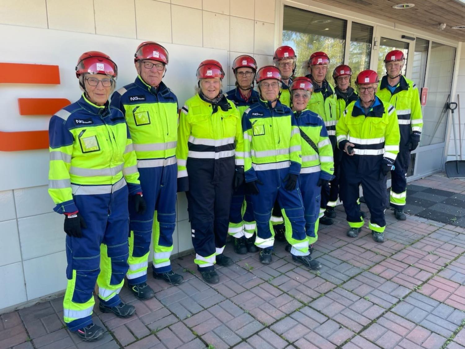 Tio personer klädda i reflexkläder och röda hjälmar står på rad utanför en byggnad och poserar för en gruppbild.