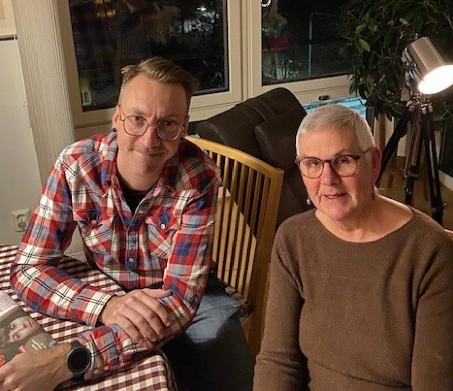 Två personer sitter vid ett rutigt bord och tittar in i kameran. En man med glasögon och rutig skjorta sitter bredvid en kvinna med kort grått hår och glasögon.