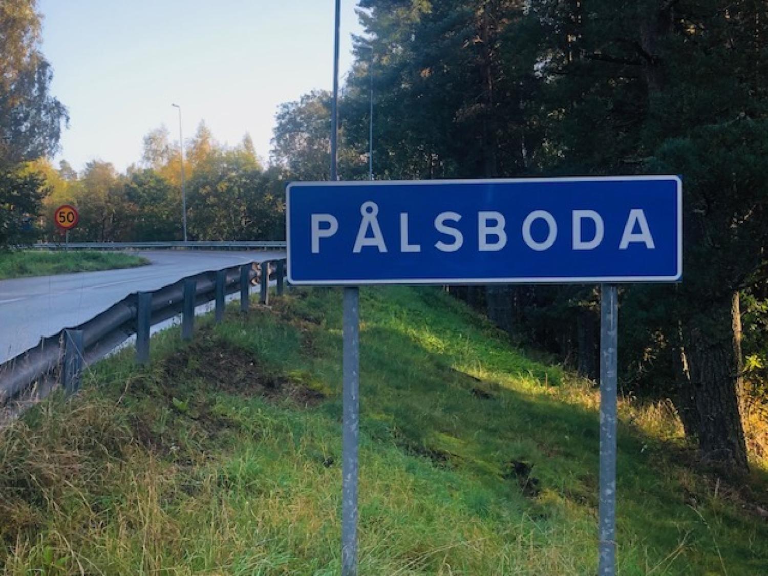 En blå vägskylt med texten ”Pålsboda” står vid sidan av en kurvig, tvåfilig väg som kantas av träd och en hastighetsskylt på 50 km/h.