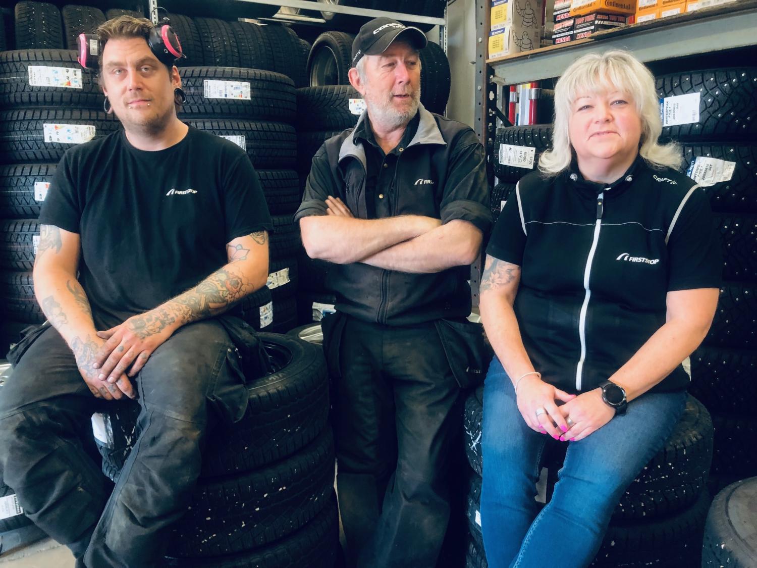 Tre personer sitter på bildäck i ett garage, omgivet av däckstaplar och hyllor med lådor. De bär matchande arbetsuniformer och ser ut att vara personal inom bilindustrin.