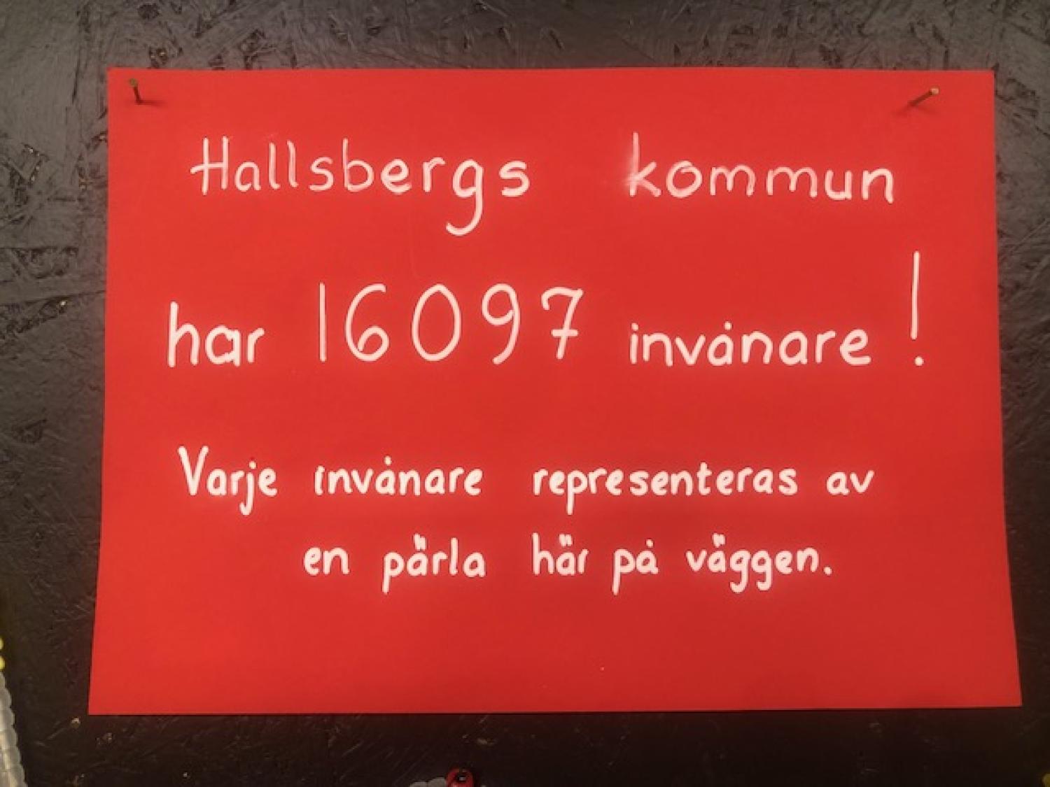 Röd skylt med vit handskriven svensk text som anger att Hallsbergs kommun har 16.097 invånare, var och en representerad av en pärla på väggen.