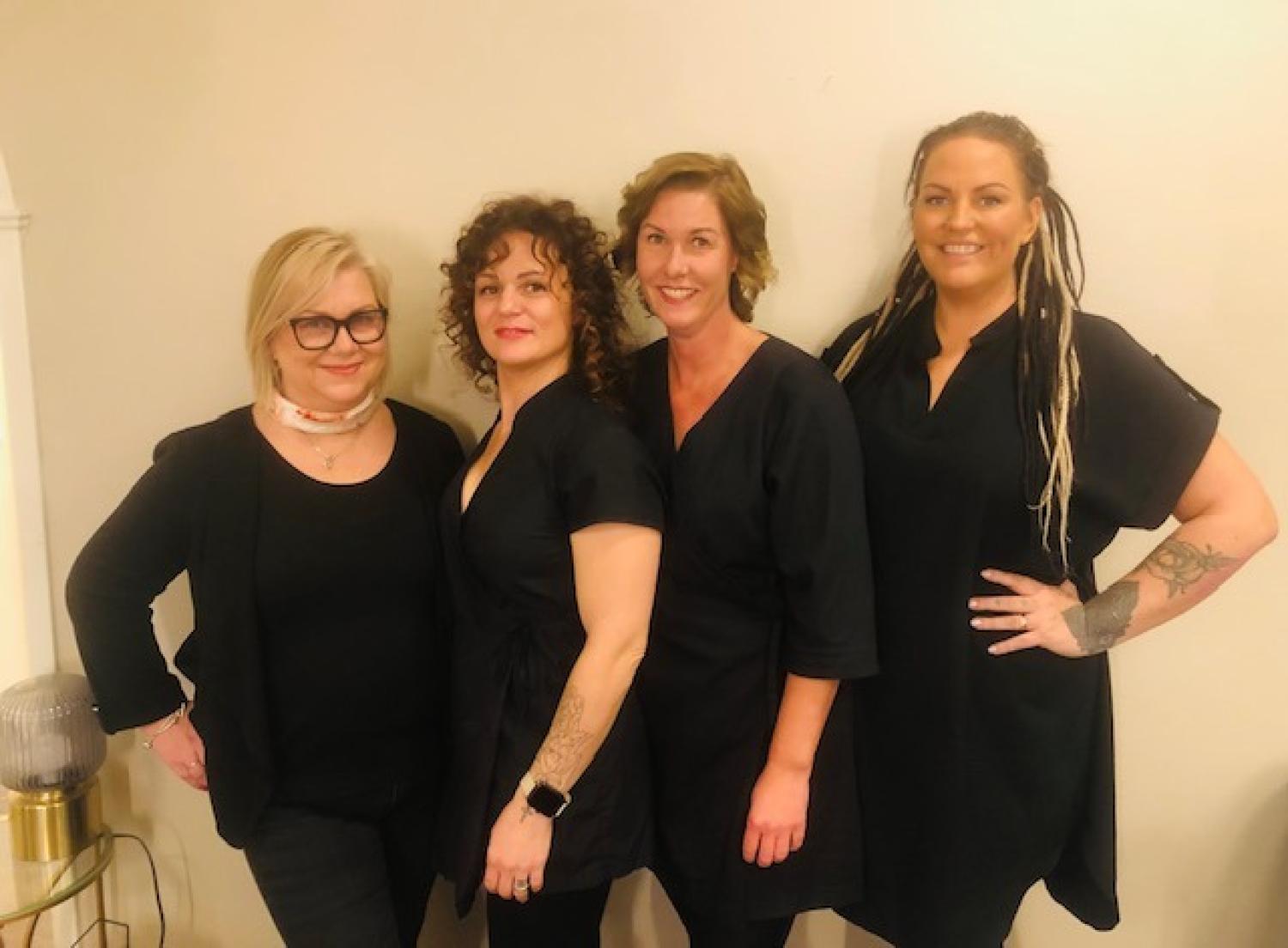 Fyra svartklädda kvinnor står sida vid sida och ler mot kameran mot en vanlig ljus vägg.