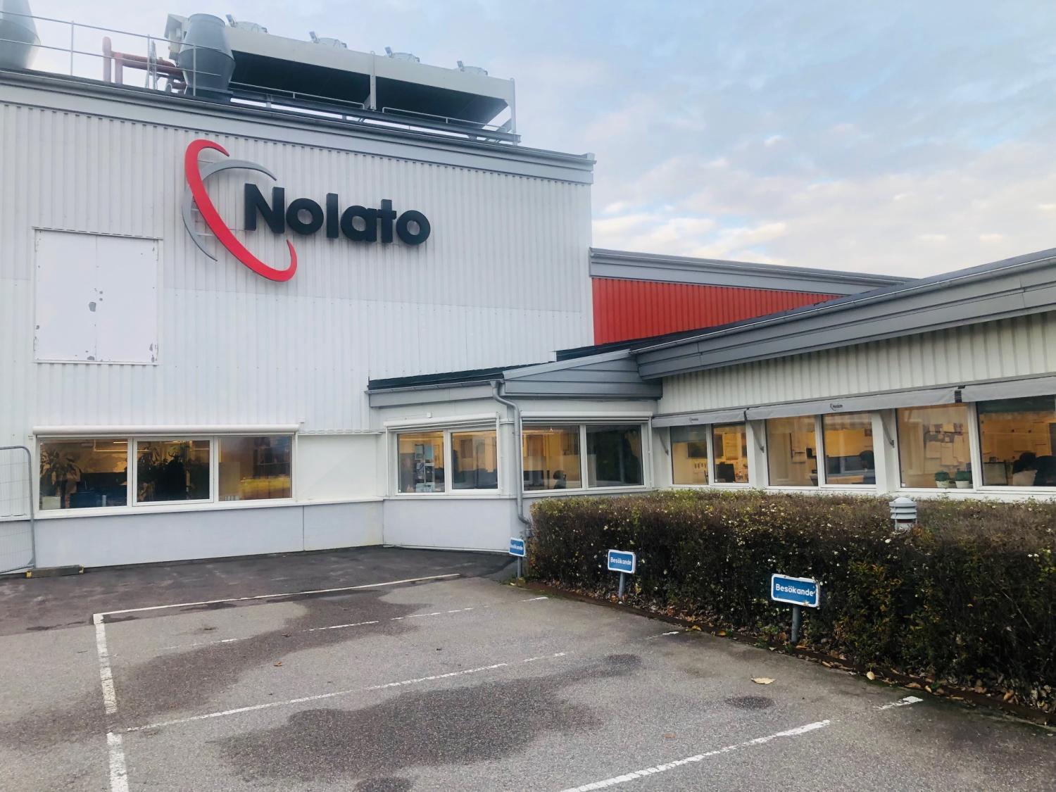 Exteriör av en Nolato-byggnad med stor logotyp, flera fönster och två reserverade parkeringsplatser på framsidan under en molnig himmel.