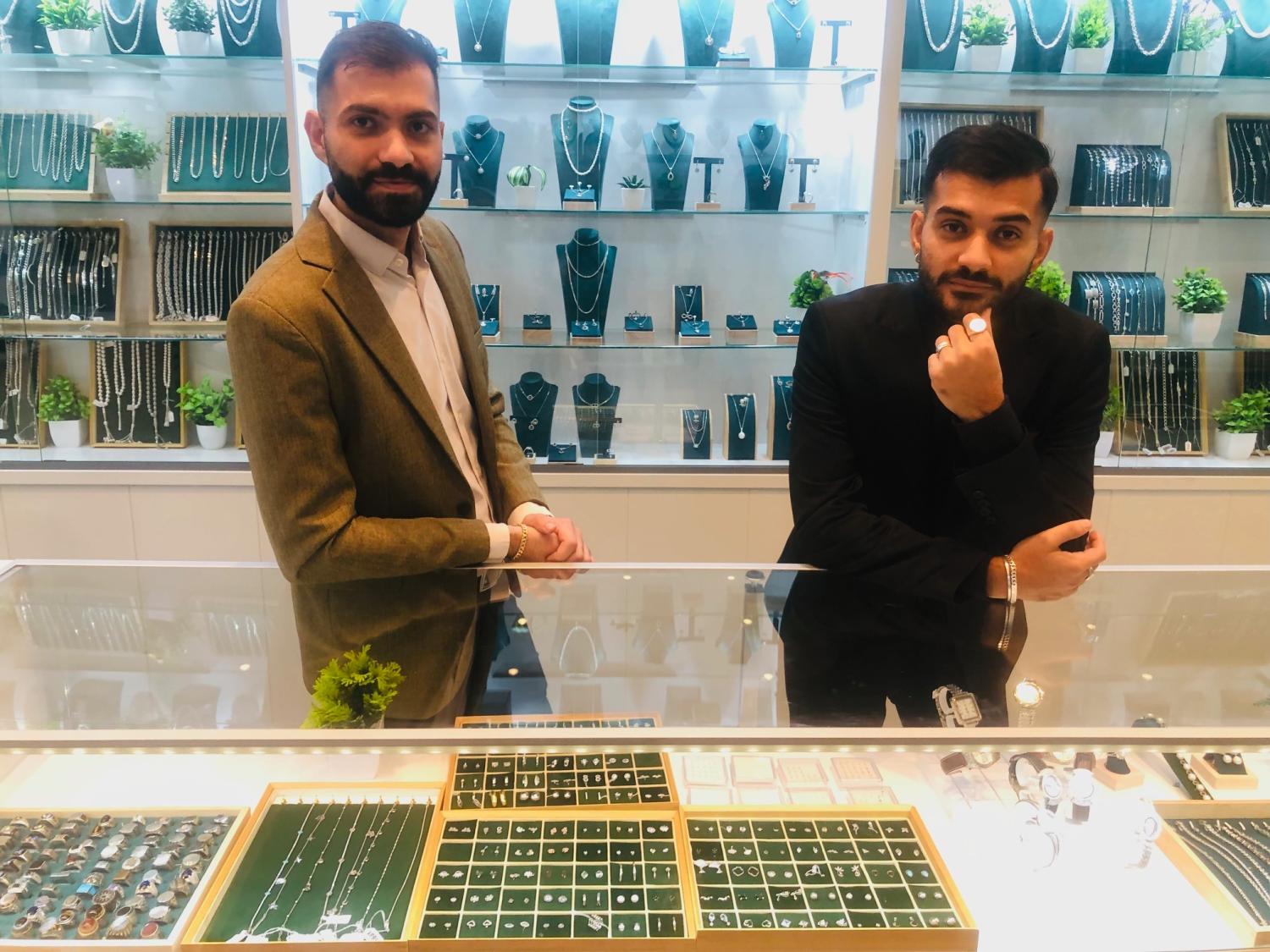 Två män står bakom en smyckesdisk i glas med ringar och accessoarer, med hyllor med halsband och smycken i bakgrunden.