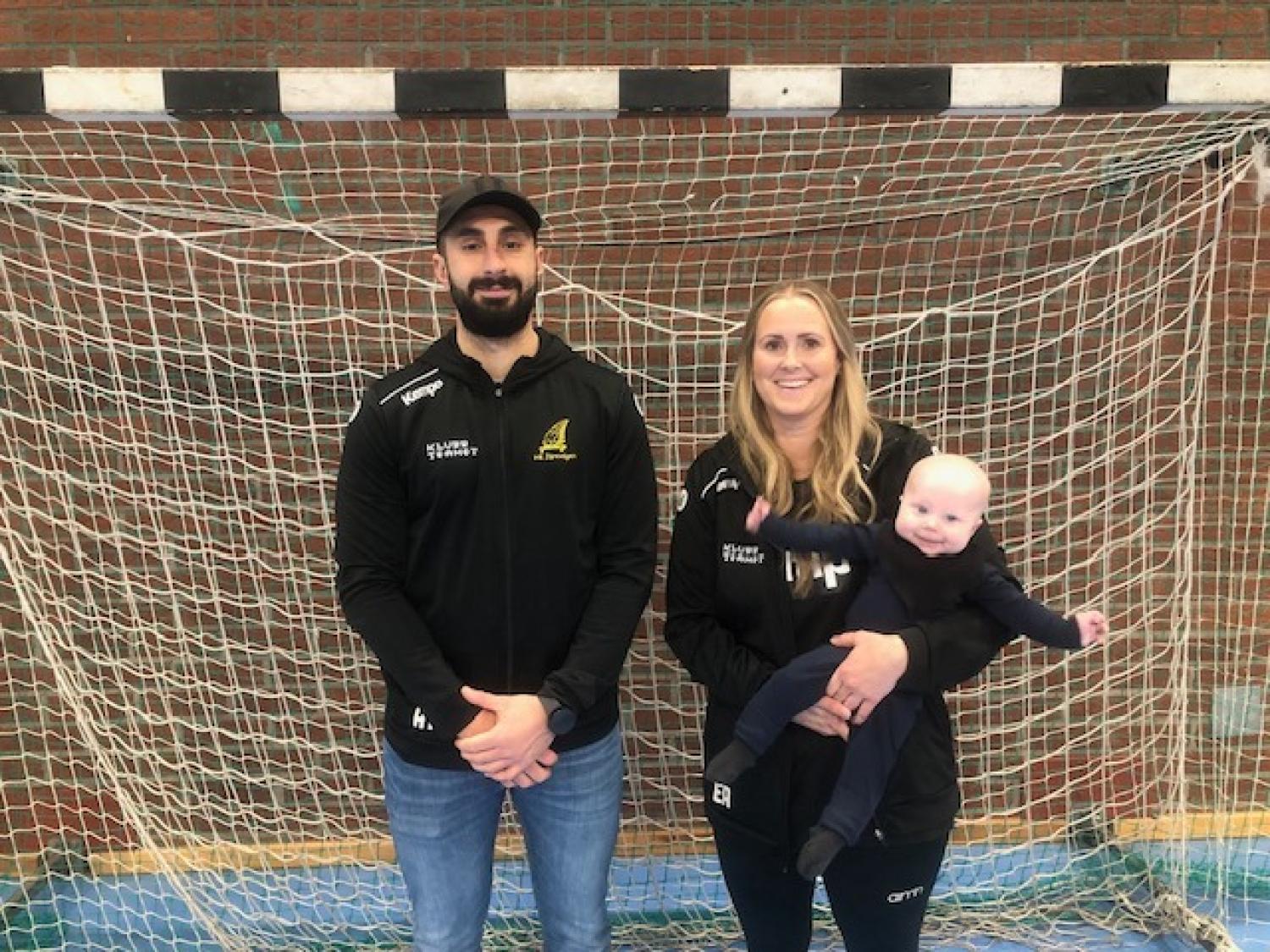 Två vuxna i sportkläder står framför ett inomhusfotbollsmål; kvinnan till höger håller i en bebis.