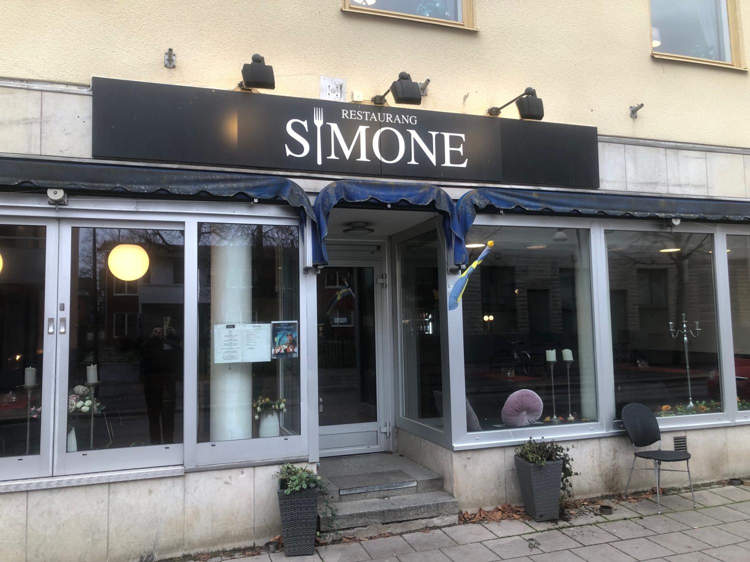 Exteriörvy av Restaurang Simone med glasfönster, meny vid dörren, svensk flagga och en ensam stol på trottoaren.