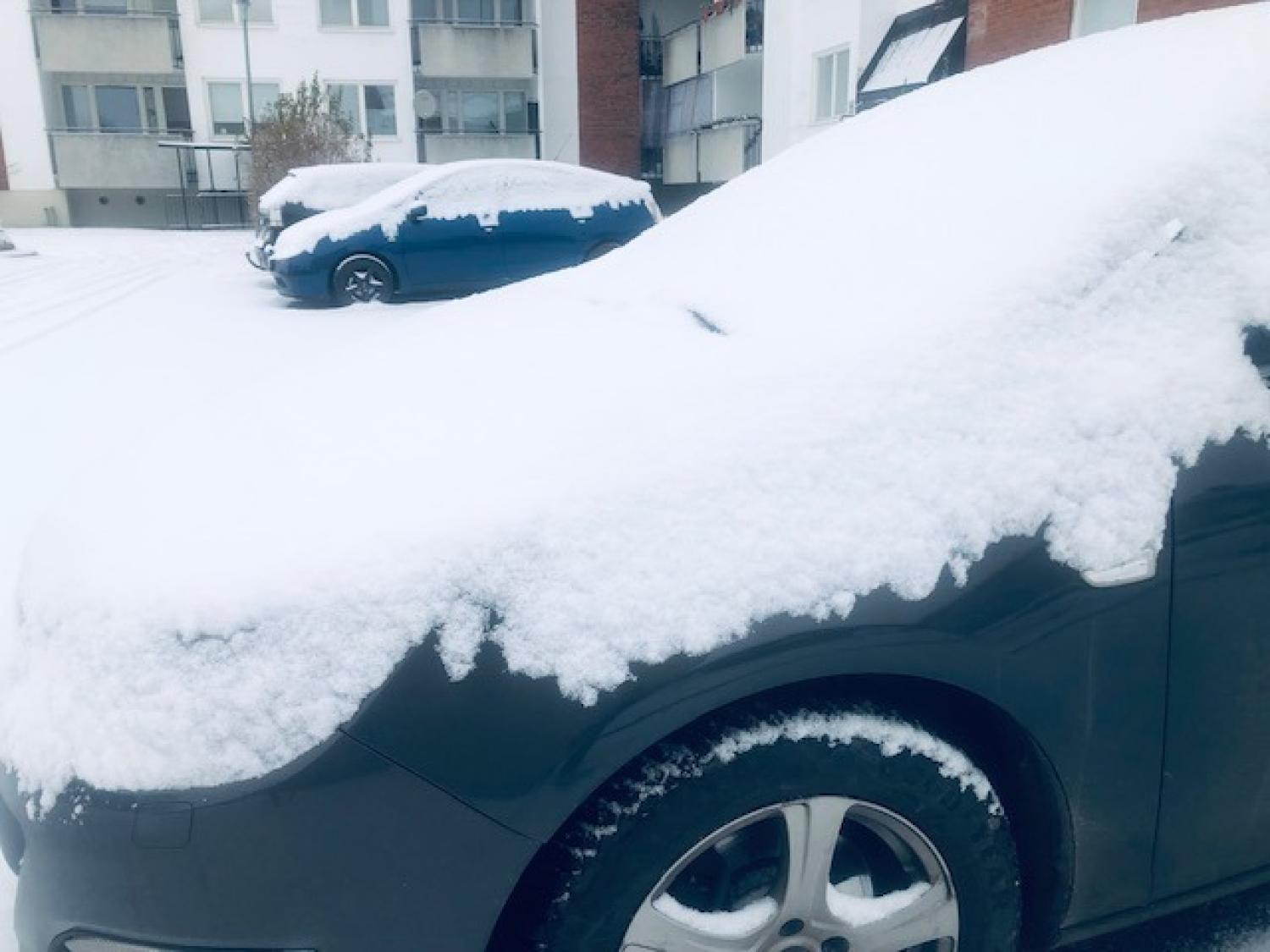 Två parkerade bilar täckta av ett lager snö på en parkeringsplats med bostadshus i bakgrunden.