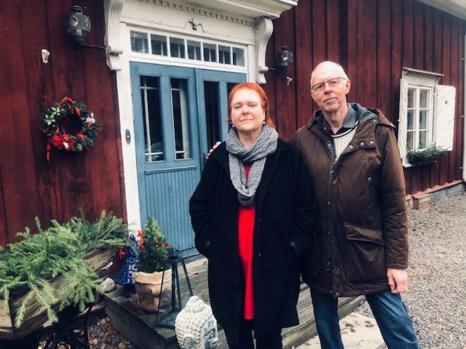 Två personer står utanför ett rött trähus med blå dubbeldörr, julkrans och vintergrönska vid entrén.