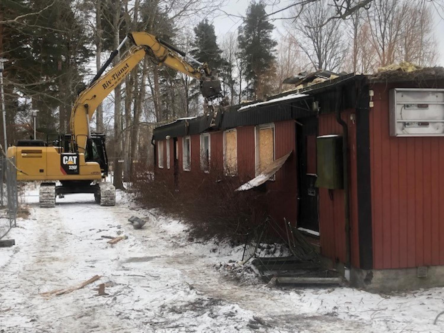 En gul grävmaskin river en röd träbyggnad på en snöig mark, med bråte utspridd i närheten och träd i bakgrunden.