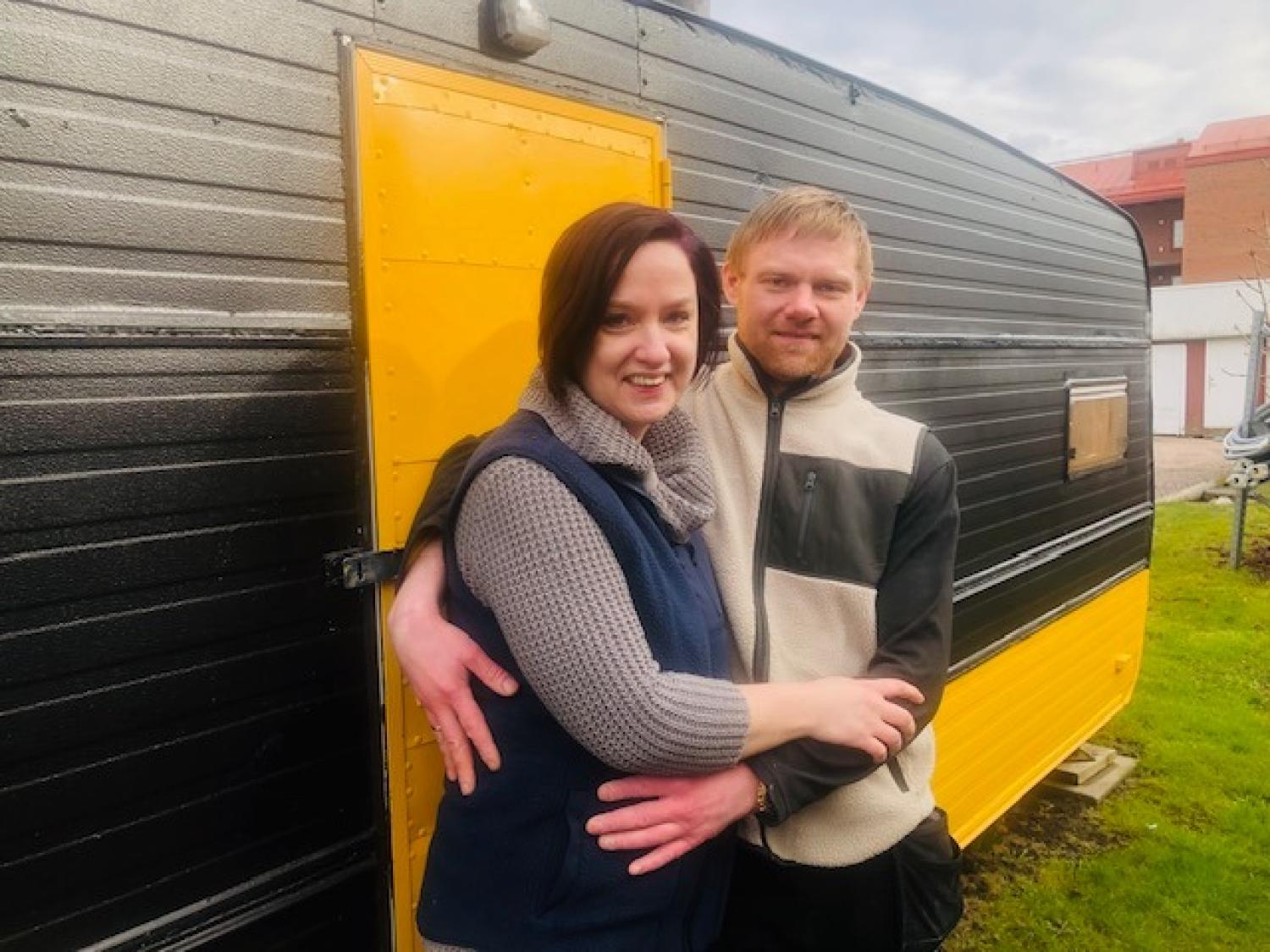 Ett par står och ler och omfamnar varandra framför en svartgul husbil på en gräsmatta.