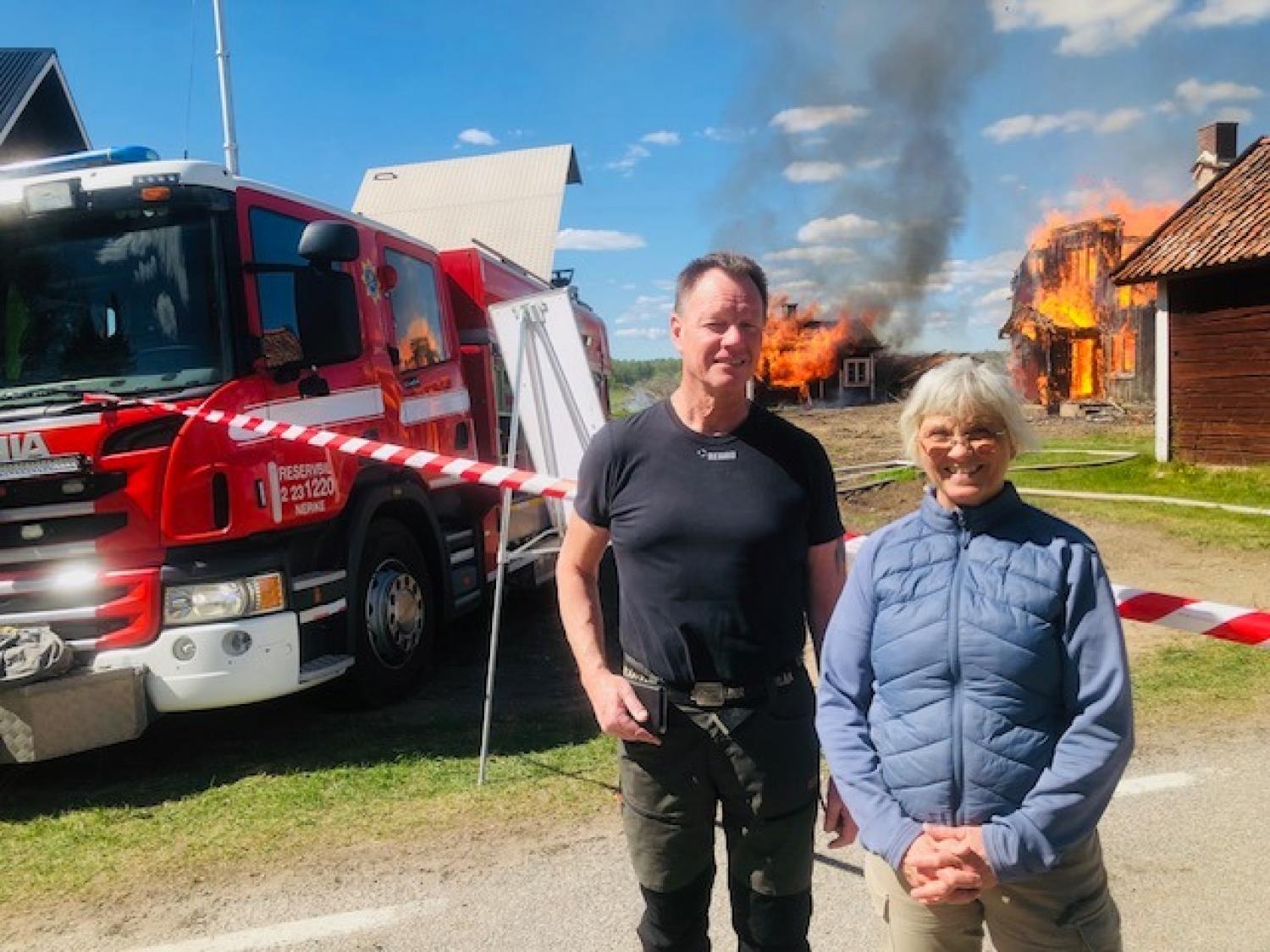 Två personer står framför en brandbil och ett avspärrat område, med ett hus som brinner i bakgrunden och rök som stiger upp mot himlen.