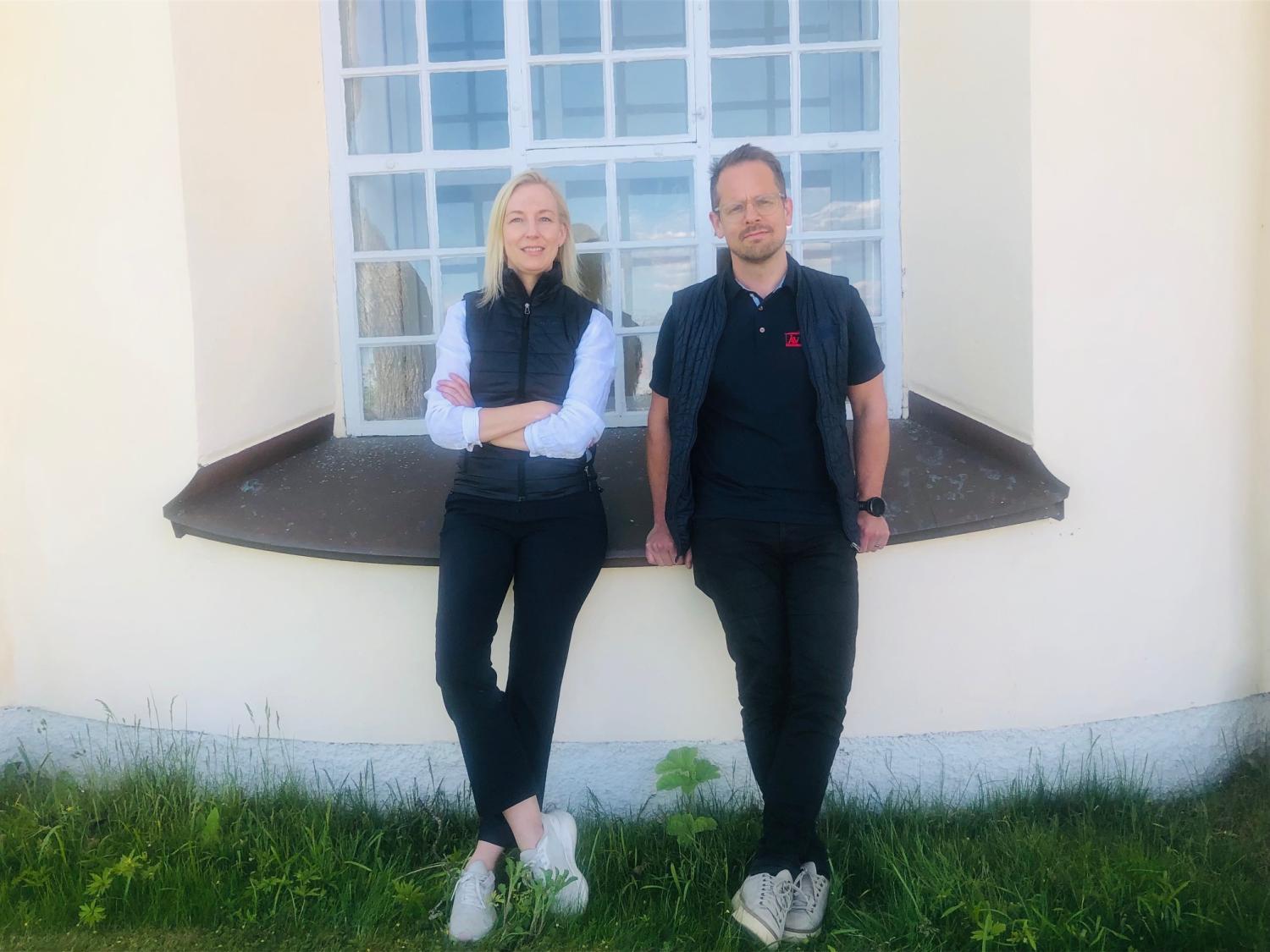 Två personer står utomhus framför ett stort fönster, lutade mot väggen med korsade armar och ben, på en gräsmatta.
