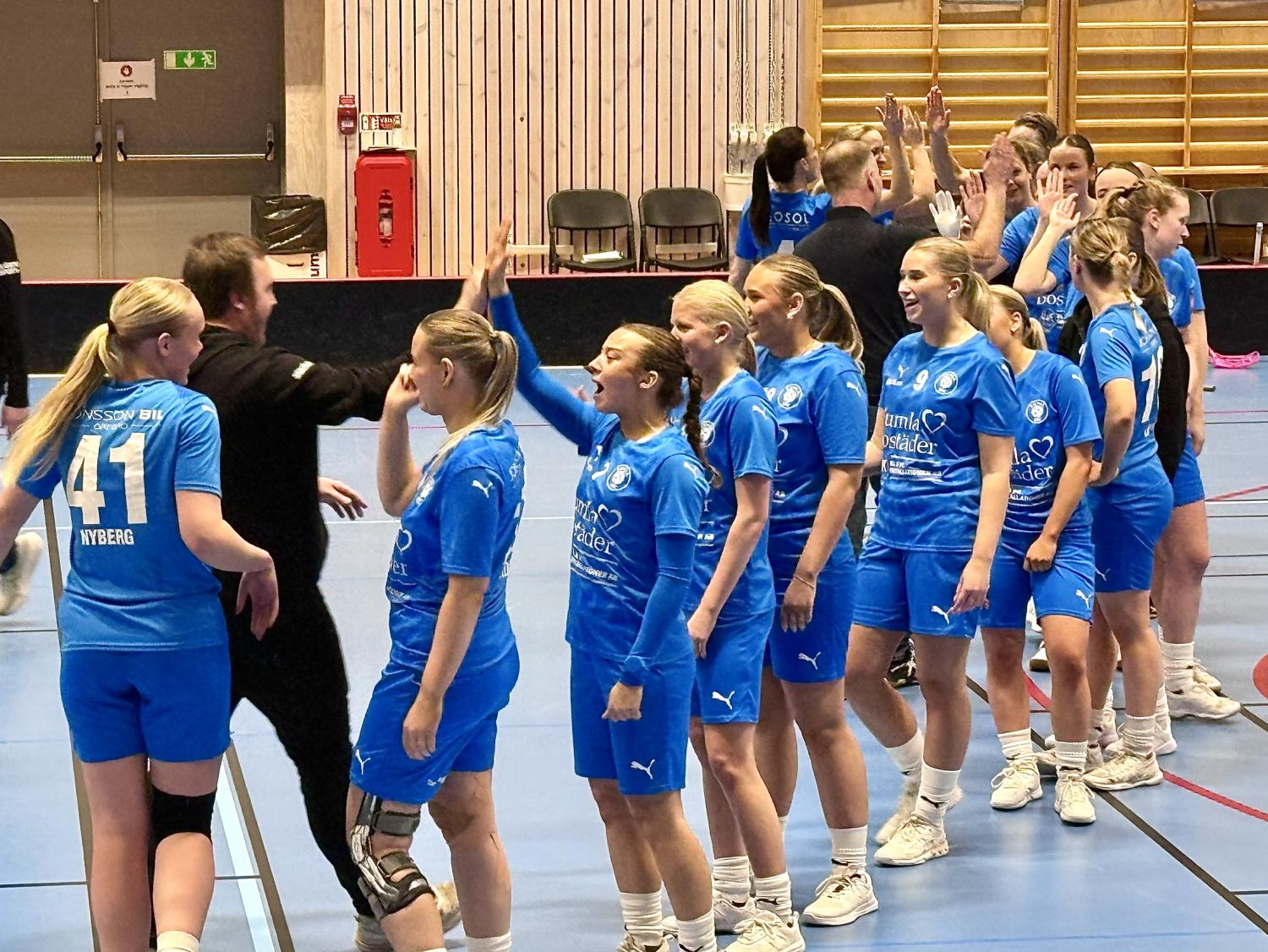Ett handbollslag för damer i blåa dräkter ställer upp sig och ger high five till tränare eller personal i en inomhushall.