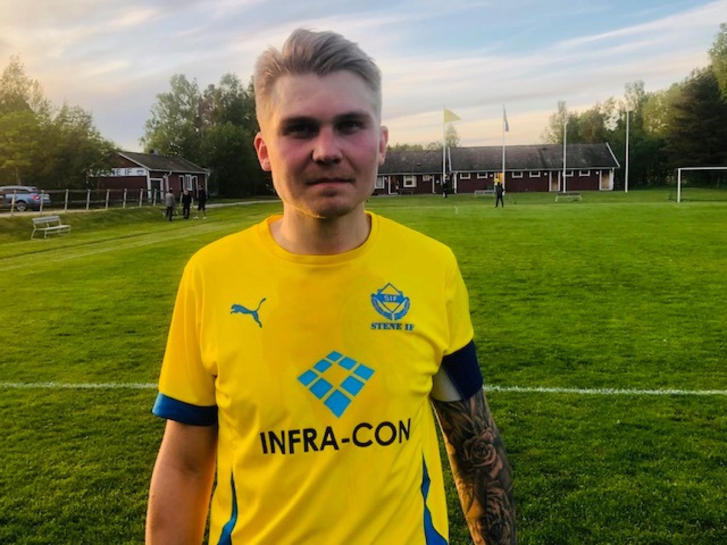 En fotbollsspelare iförd en gul Stensele IF-tröja står på en grön plan med träd och byggnader i bakgrunden.