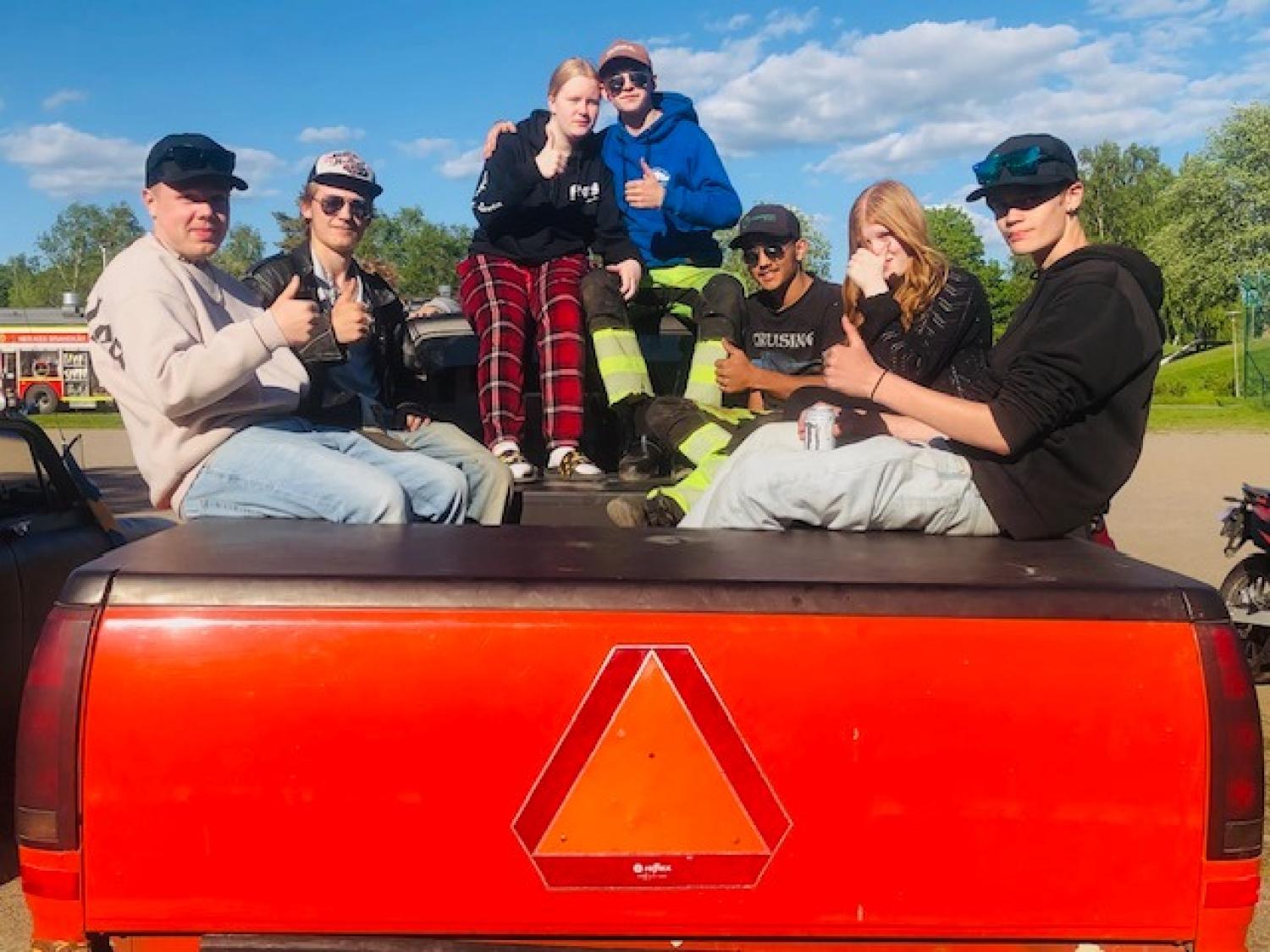 Sju personer sitter på baksidan av ett rött fordon, gör tummen upp och poserar för kameran en solig dag. Träd och en blå himmel syns i bakgrunden.