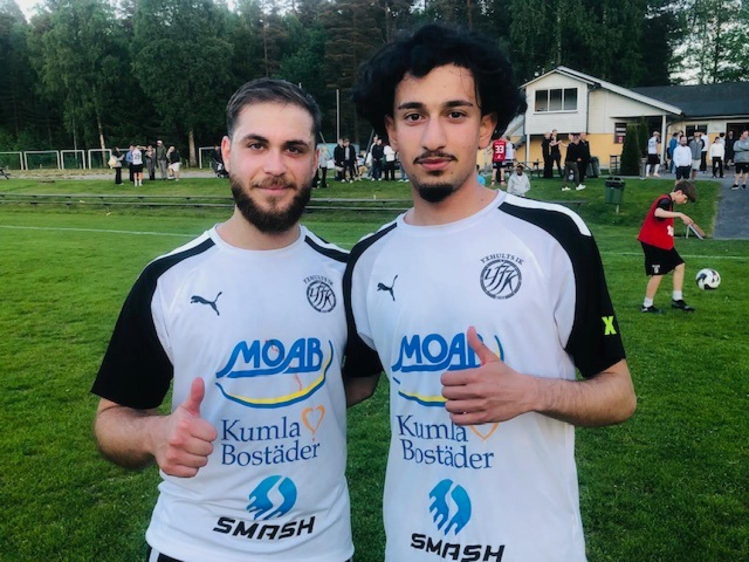 Två fotbollsspelare i vita tröjor står på en gräsplan och gör tummen upp, med människor och en byggnad i bakgrunden.