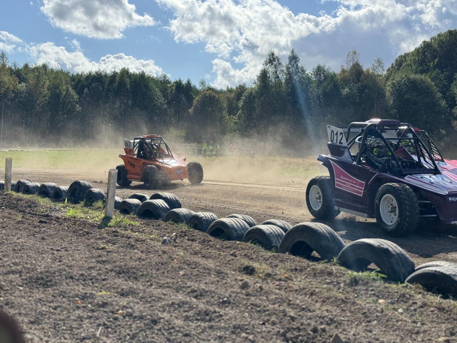 Två terrängbilar tävlar på en grusbana och sparkar upp damm, med ett skogsområde och en delvis molnig himmel i bakgrunden.